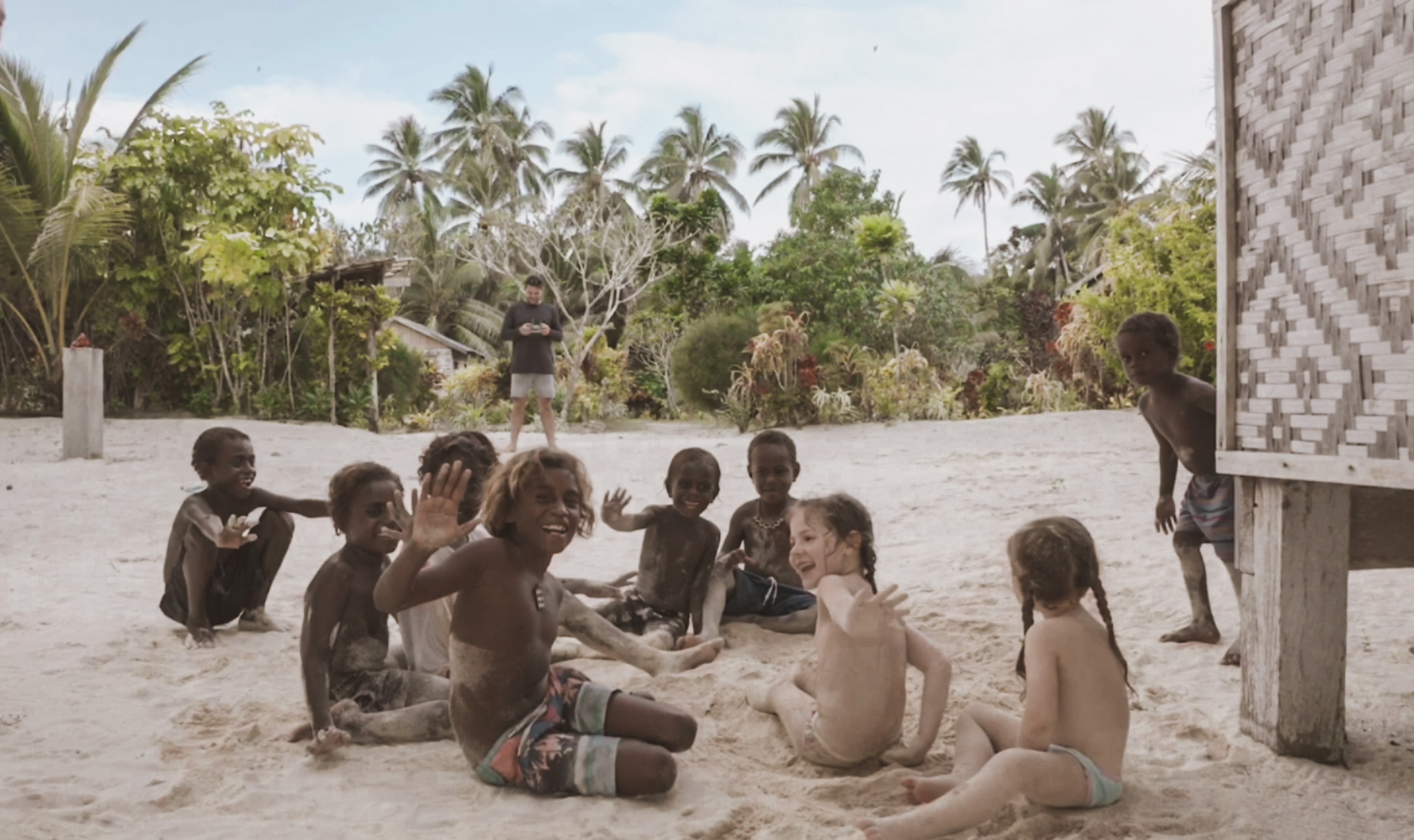 Group of children, including two white Australian girls and several local dark-skinned children, sitting together on the beach next to a hut in the Solomon Islands, laughing and waving. This scene captures the importance of a family vacation by demonstrating how travel fosters friendships, cultural exchange, and mutual understanding among children from diverse backgrounds.