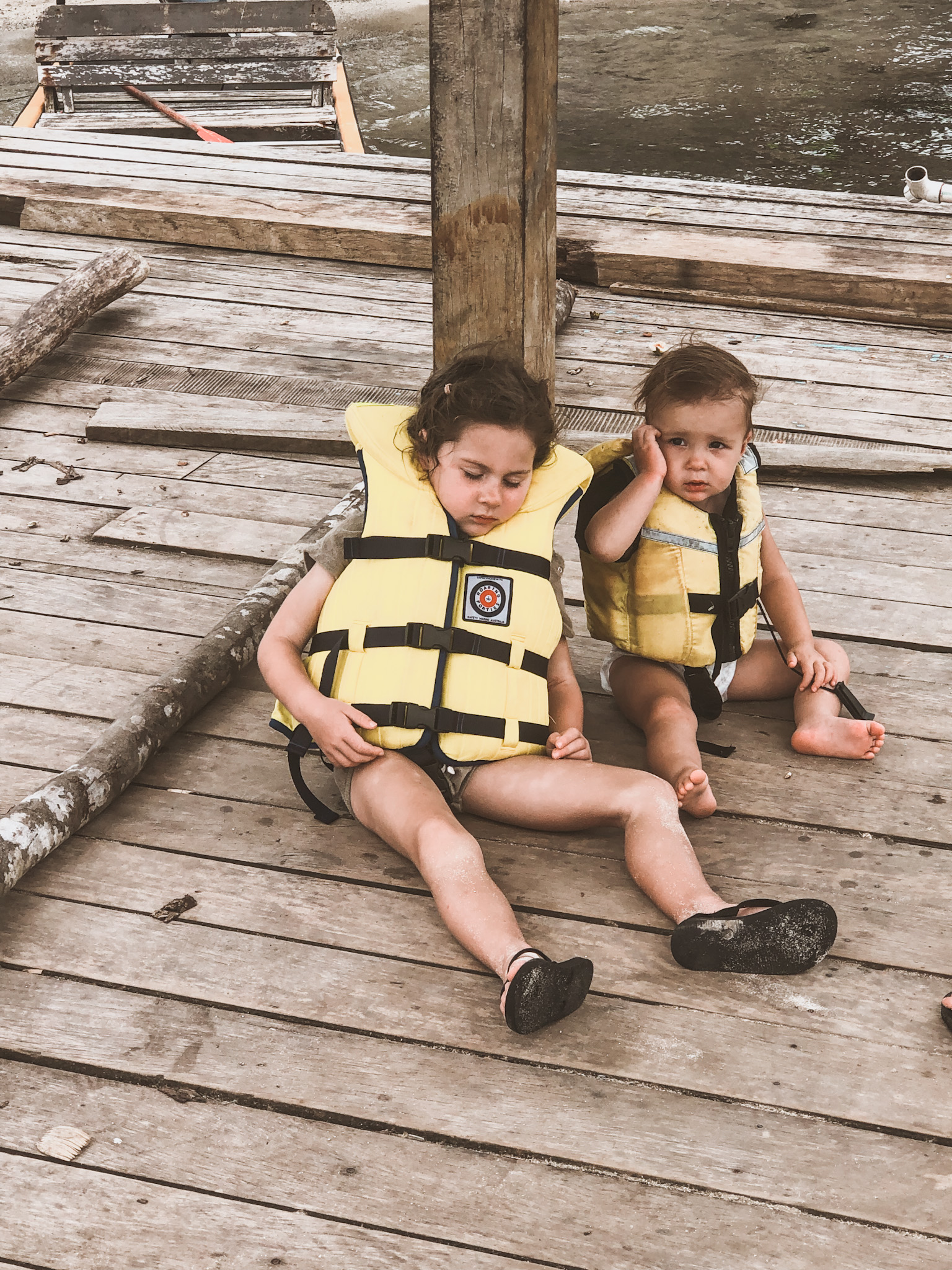 Two young sisters on a jetty after a boat trip, still in their life jackets with one girl asleep, symbolizing the importance of a family vacation by showing how travel with children encourages parents to embrace slower moments together.