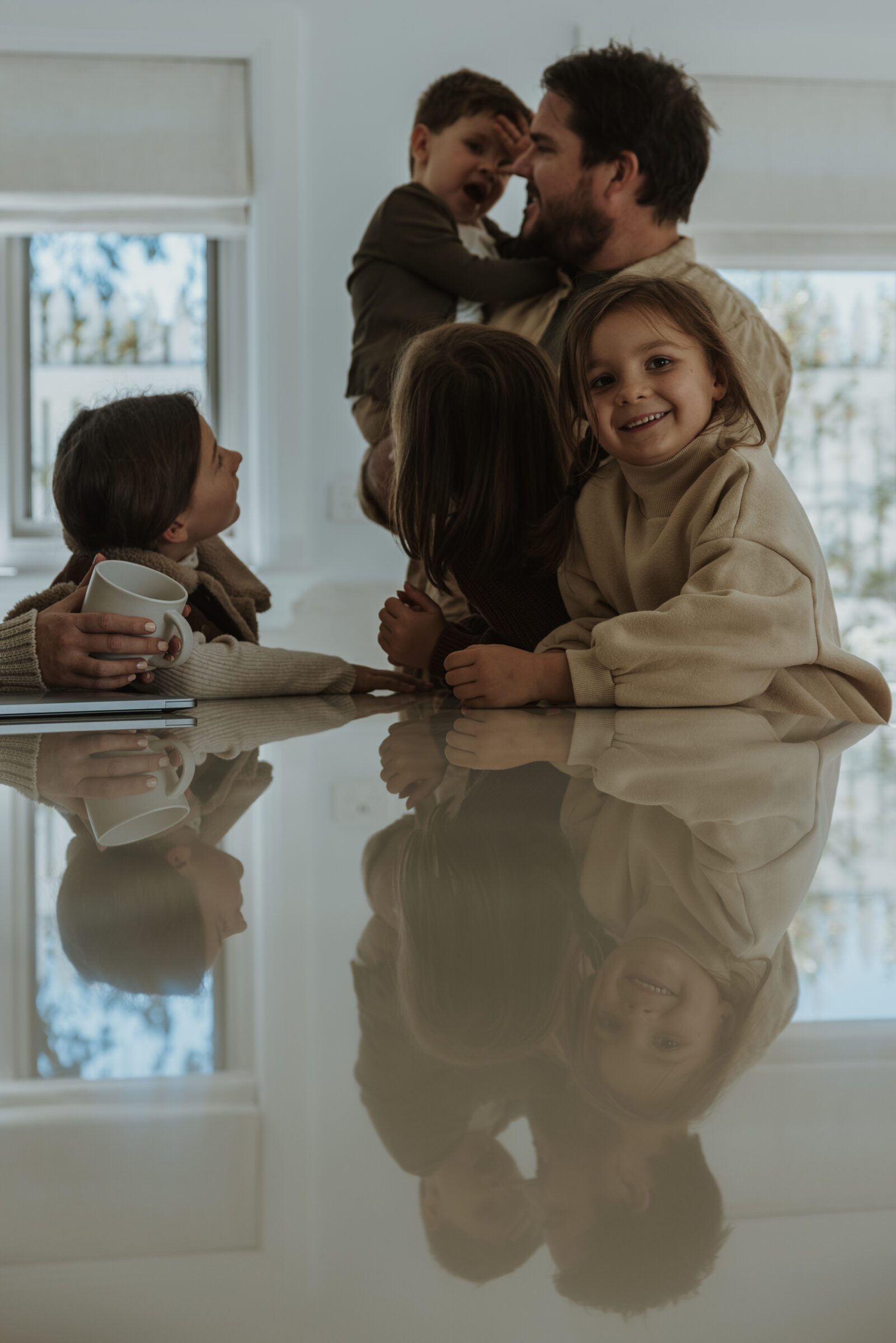 Father holding his son with three daughters gathered around the kitchen benchtop, discussing preparations for their airport departure, depicting the family involvement in planning for a family vacation, highlighting the importance of communication and coordination before setting off.