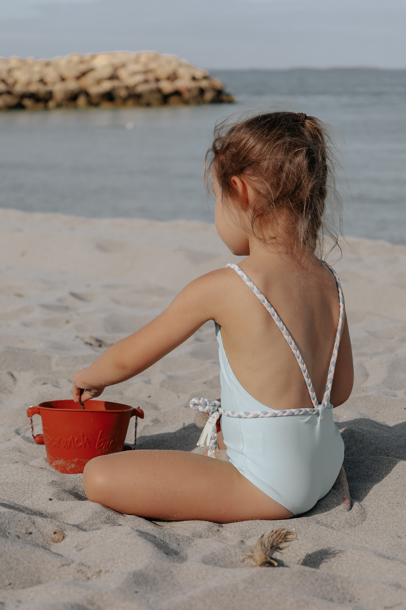 Young girl sitting in the sand on the beach, playing with a stick and a sand bucket, symbolizing the timeless appeal of beach vacations as a winning choice for family destinations, embodying simplicity and joy in deciding where to take a family vacation.