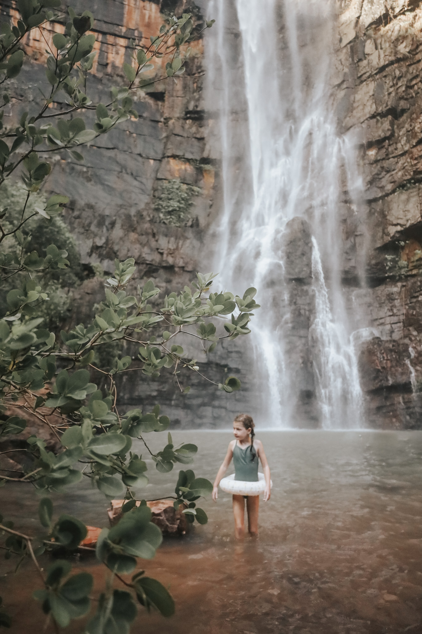 Young girl walking in a natural swimming hole with a giant waterfall in the Australian Kimberley during the wet season, showcasing how to plan for a family vacation by taking advantage of off-season travel for cheaper flights and unique experiences, like enjoying the full flow of waterfalls.