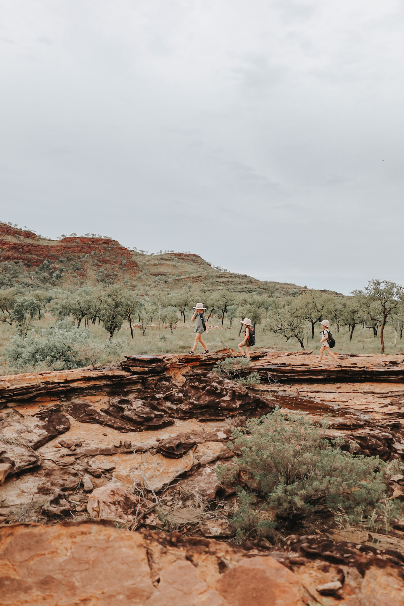 Three young sisters hiking in the national parks of Kununurra, Western Australia, with the vibrant orange and green landscape of the Australian Kimberley in the background, representing a wonderful family holiday destination in Australia and showcasing the natural beauty and adventure ideal for family vacations.