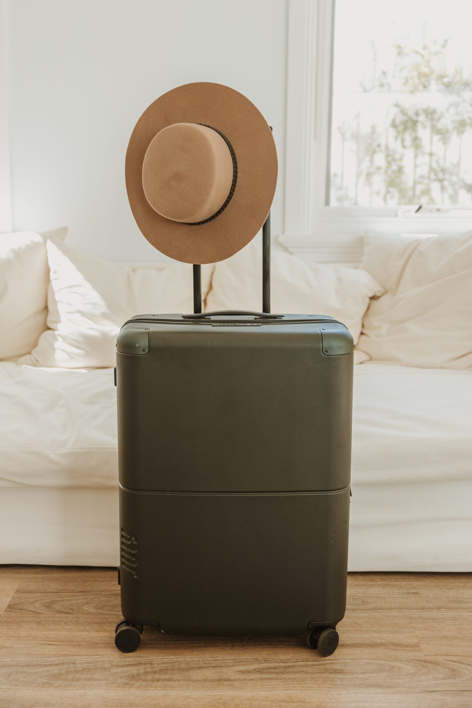 No problem! Here's the revised alt text with the correct keyword: Dark green suitcase in front of a white couch with a light brown wide brim hat hanging off its handle, showcasing efficient packing for traveling with kids and emphasizing the importance of knowing airline baggage restrictions to ease airport stress.