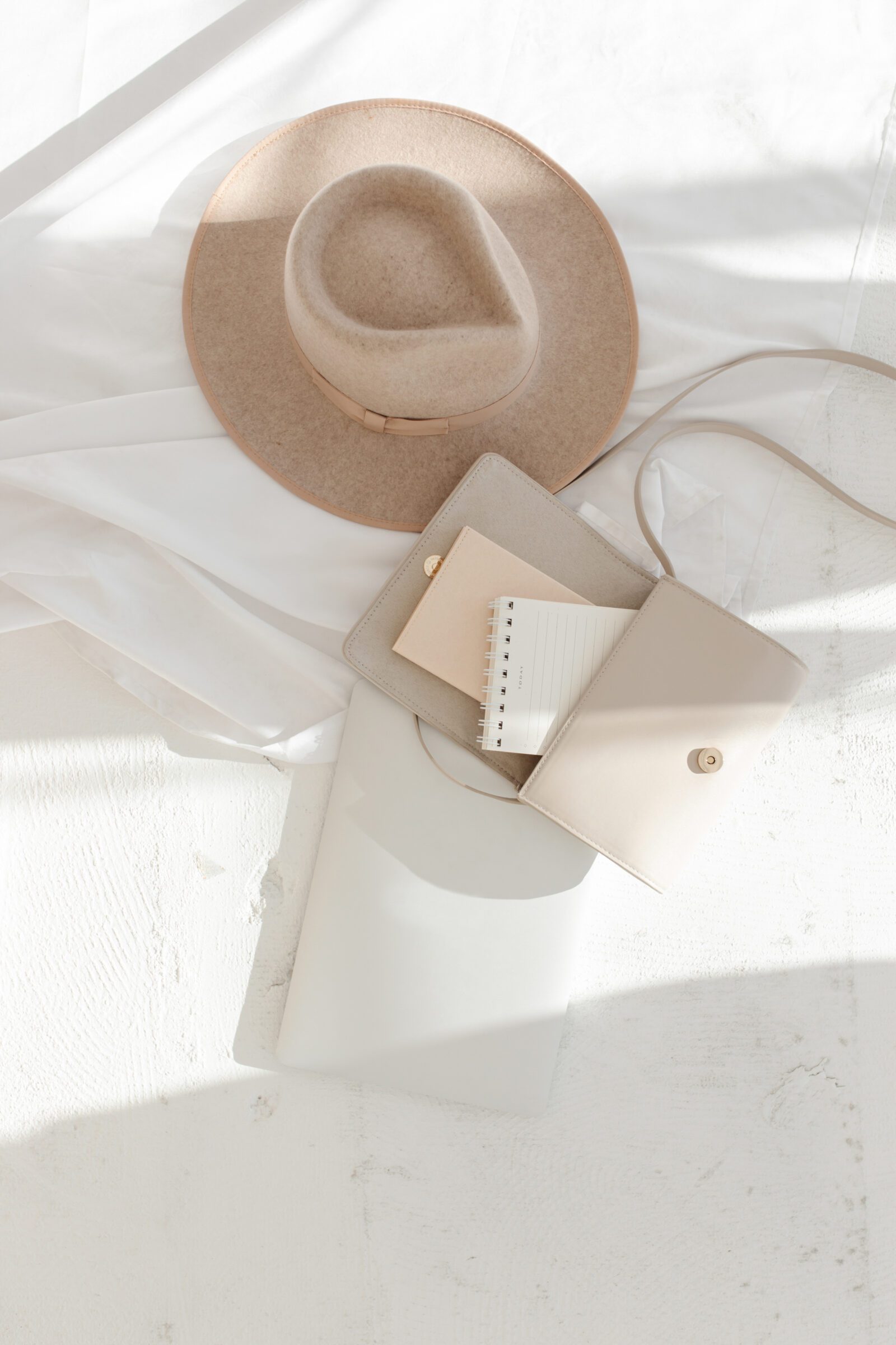 Woman's travel essentials laid out on a white textured background, including a hat, an open beige purse with a notebook and small wallet inside, and a laptop, illustrating the best way to bring money on a trip through careful planning and organization.