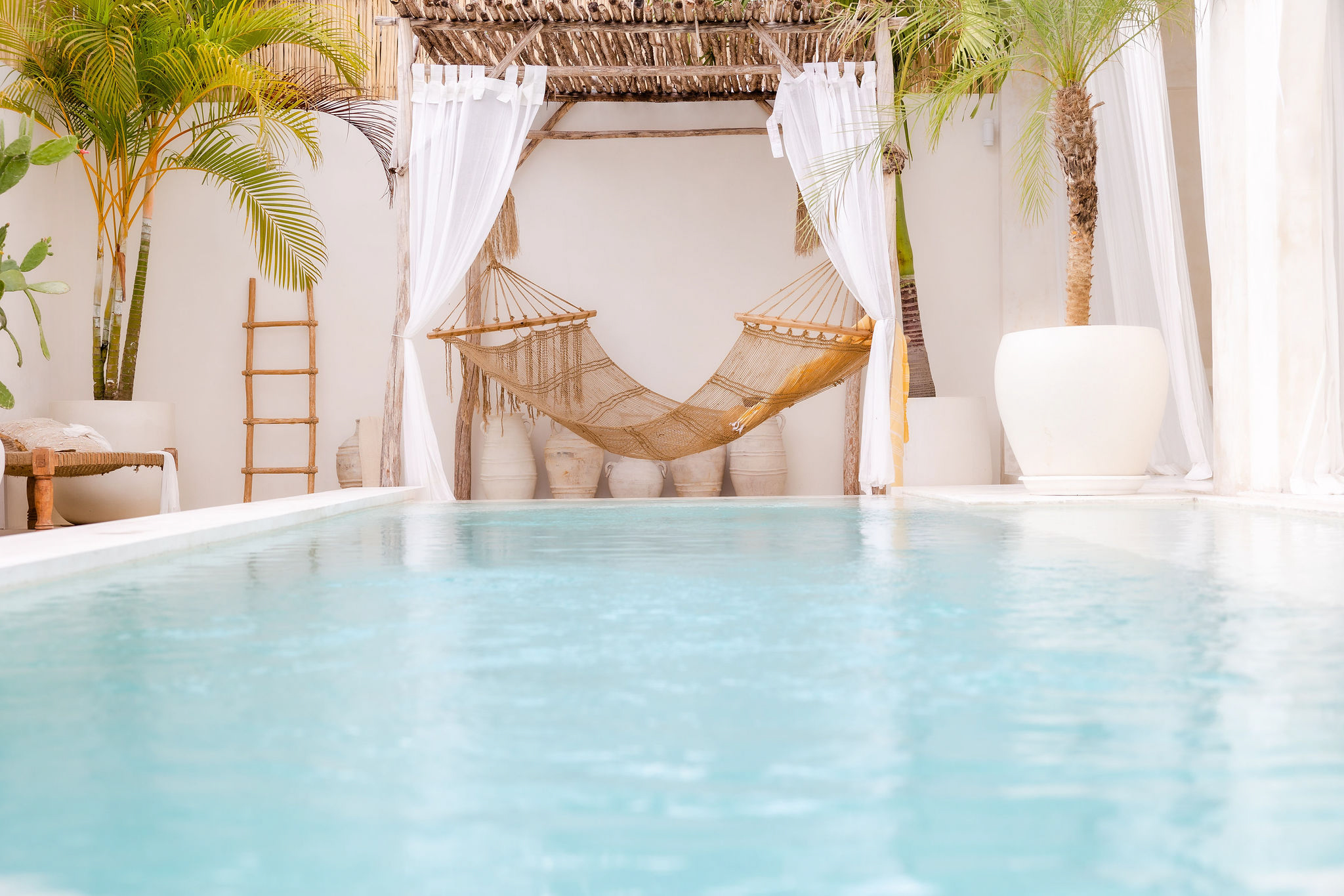 Hotel private pool area, framed by white walls and curtains, adorned with white towels and small potted palm trees, featuring a wooden cabana with a stylish brown hammock, encapsulating high-low travel by showcasing luxurious accommodation that has been prioritised within a family's travel budget.