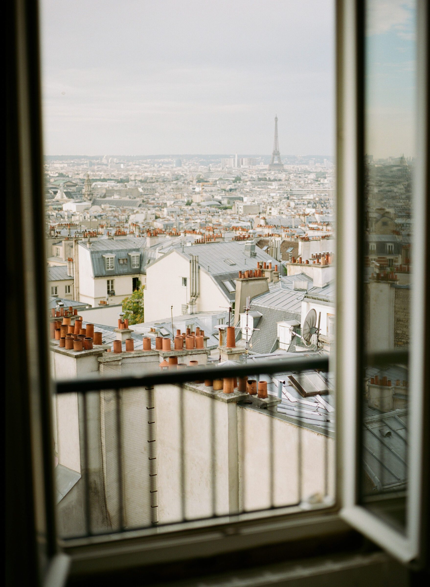 Eiffel Tower seen through the window of a hotel room, with Parisian rooftops in the foreground, exemplifying the splurge on accommodation with a view as part of the high-low travel method, blending premium experiences with budget-conscious planning.