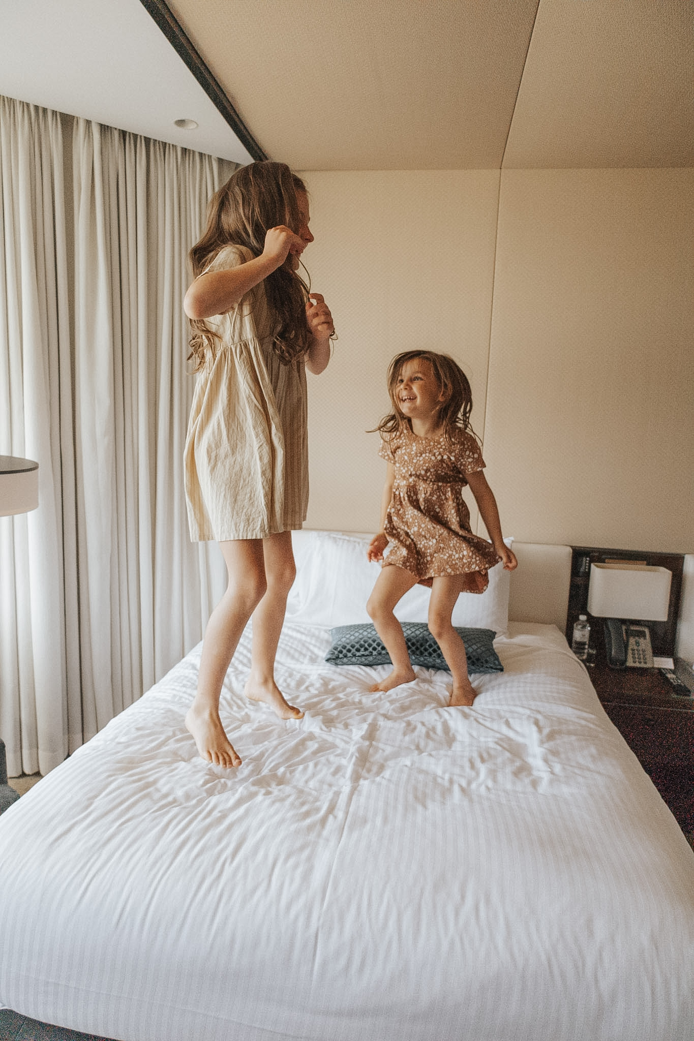 Two young sisters jumping on a hotel bed, exemplifying the joy of family vacations and underscoring accommodation as one of the largest expenses, emphasizing the importance of early booking in planning for a family vacation to secure the best deals and ensure memorable experiences.