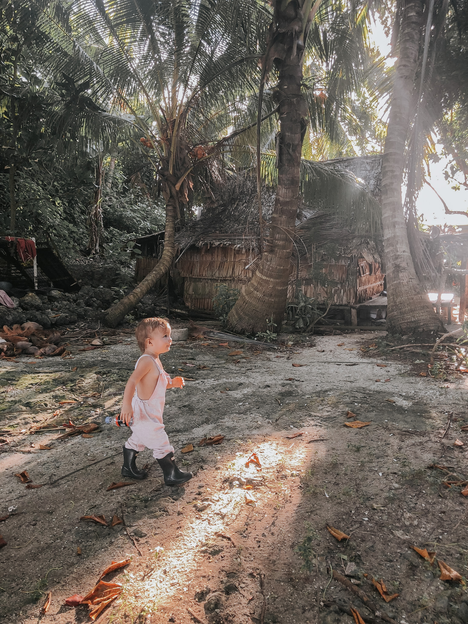 Young toddler walking in gumboots on an island in the Solomon Islands, with a wooden hut and palm trees in the background, encapsulating the considerations in planning a family vacation, such as kid-friendliness, safety, freedom for exploration, and cultural immersion, highlighting the need to tailor vacation destinations to family preferences and requirements.