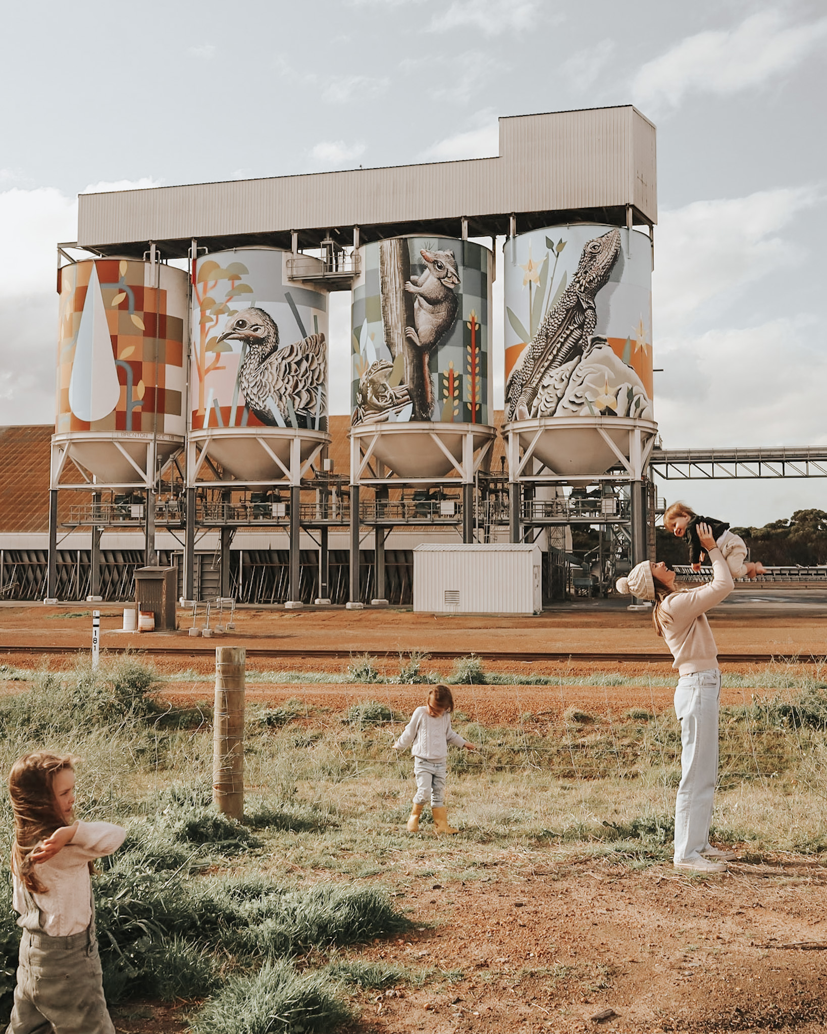 Mother lifting her baby high with joy, while two girls explore nearby, set against a rural Australian backdrop with artistically painted silo bins, highlighting the importance of a family vacation in nurturing exploration and bonding.