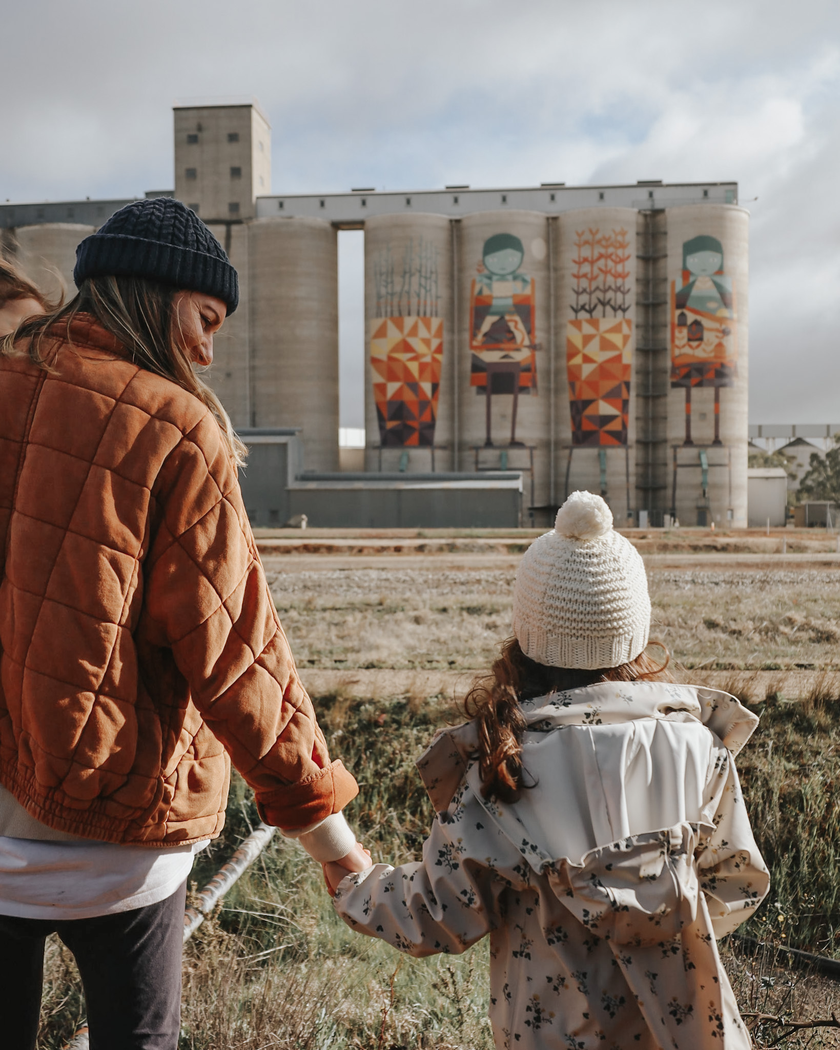 Mother and daughter, hand in hand, gazing at uniquely painted silo bins in rural Australia, embodying the joy and planning involved in affording your next family vacation.