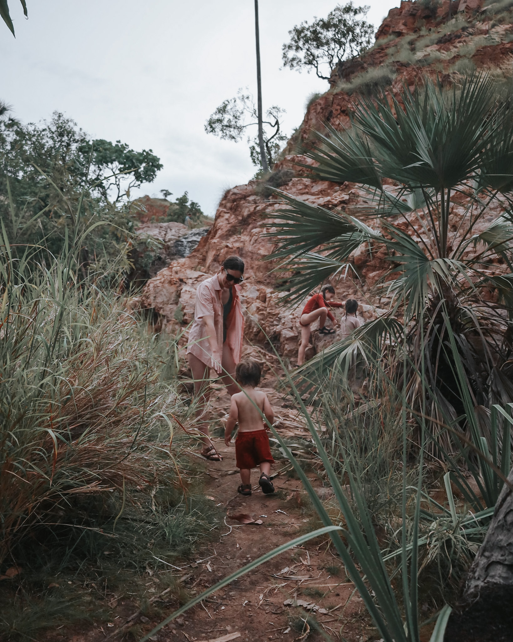 Mother guiding her toddler through bushland, with older sisters climbing rocks behind, highlighting family vacation planning that caters to all ages, ensuring activities and destinations meet everyone's needs.