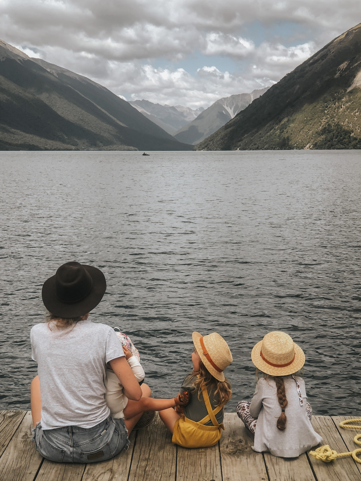 Three young girls and their mother sitting on a jetty, gazing out at a lake and mountains in New Zealand, exemplifying a fantastic overseas destination for families and illustrating the thoughtful process of deciding where to take a family vacation.