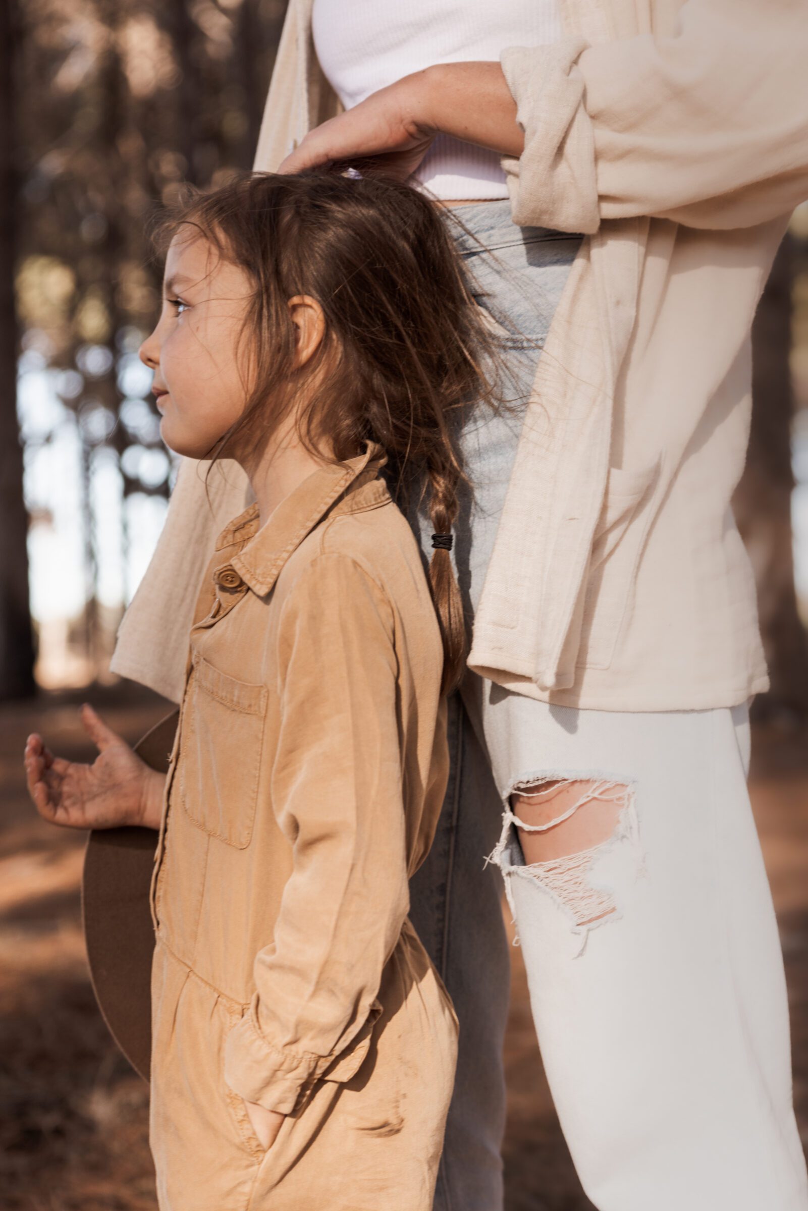 Young girl standing in front of her mother in the forest, looking happy as the mother strokes her daughter's hair, embodying the joy of family adventures and answering the question, 'Does travel have to stop once you have kids?' with a resounding no, showcasing the continued exploration and bonding experiences travel offers families.