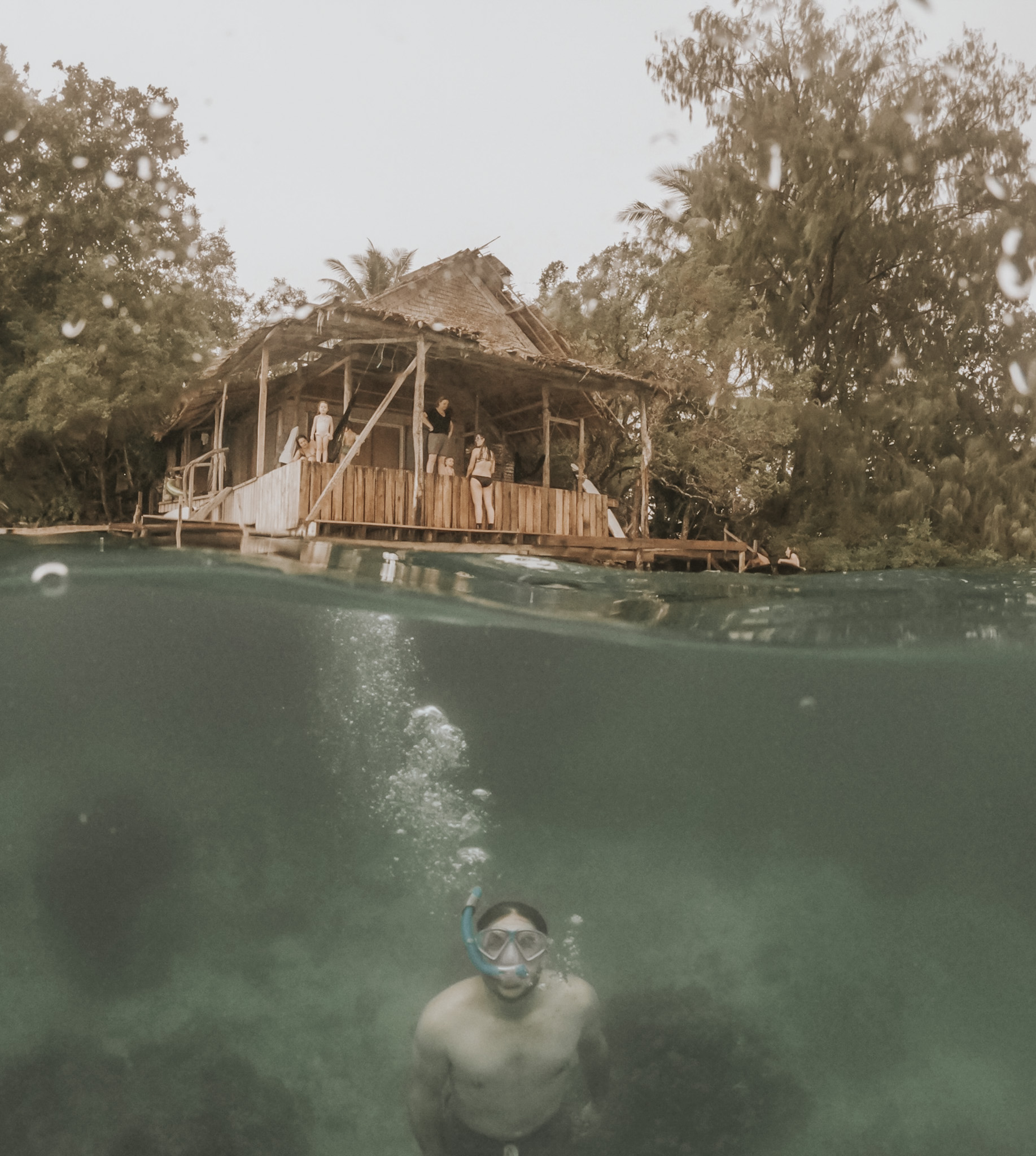 Overwater bungalow in the Solomon Islands with a family on the balcony and a man swimming below, embodying the 'high' in high-low travel by integrating a luxury stay into a balanced travel strategy that mixes indulgent experiences with budget-friendly activities.