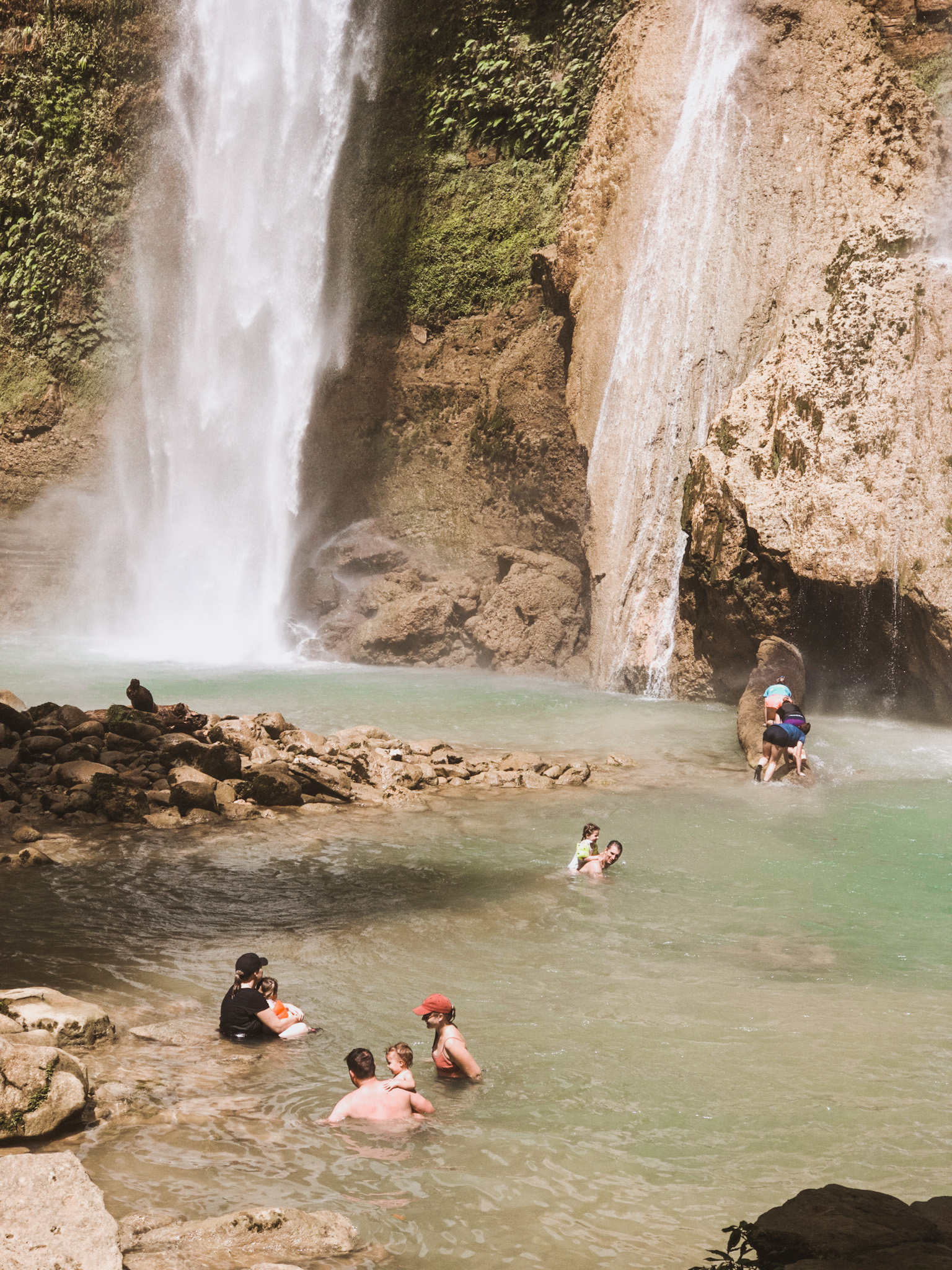 Extended family swimming together in a natural water hole under a giant waterfall in the Solomon Islands, including uncles, aunties, and cousins, showcasing the value of choosing vacation destinations that offer memorable bonding experiences for the whole family.