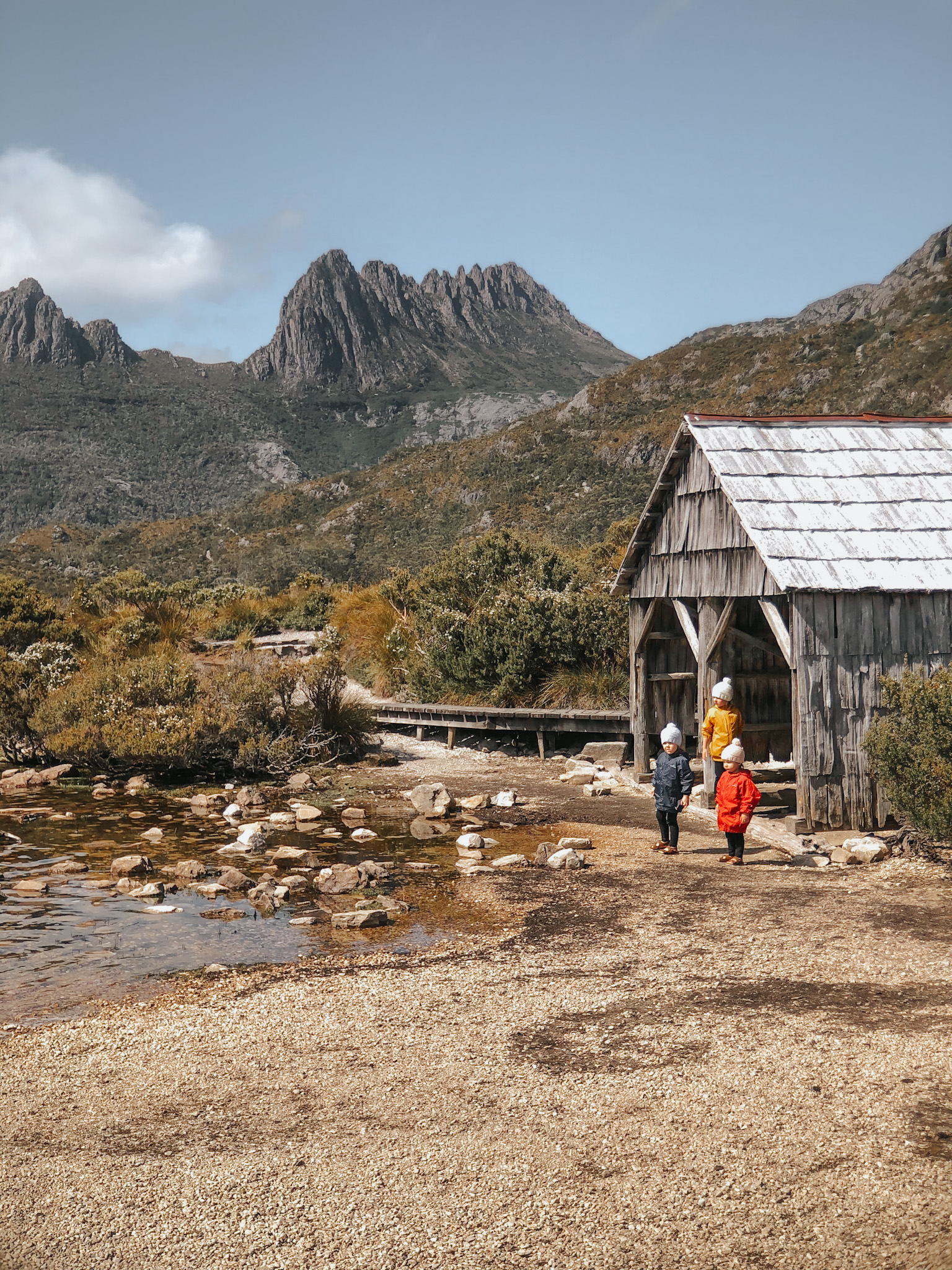 Three young sisters standing by a lake with Cradle Mountain in the background in Tasmania, during a family hike, showcasing a nature destination as one possible choice for families implementing high-low travel, based on their specific vacation goals and interests.