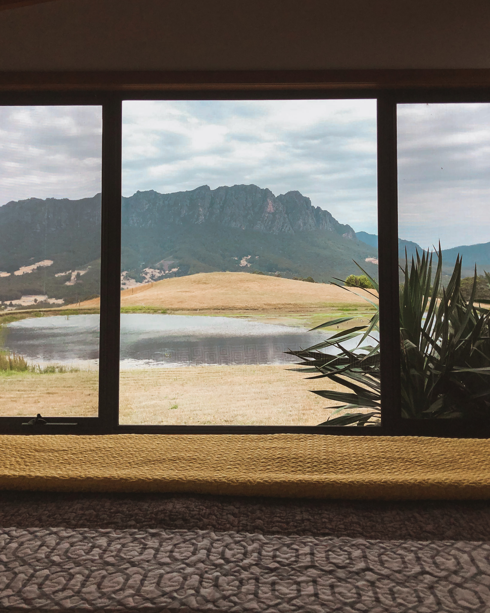 End of a luxurious bed leading to a window with a breathtaking view of Tasmanian mountains and a lake, illustrating high-low travel by combining high-end accommodation with the authentic, low-cost activity of enjoying nature's beauty.