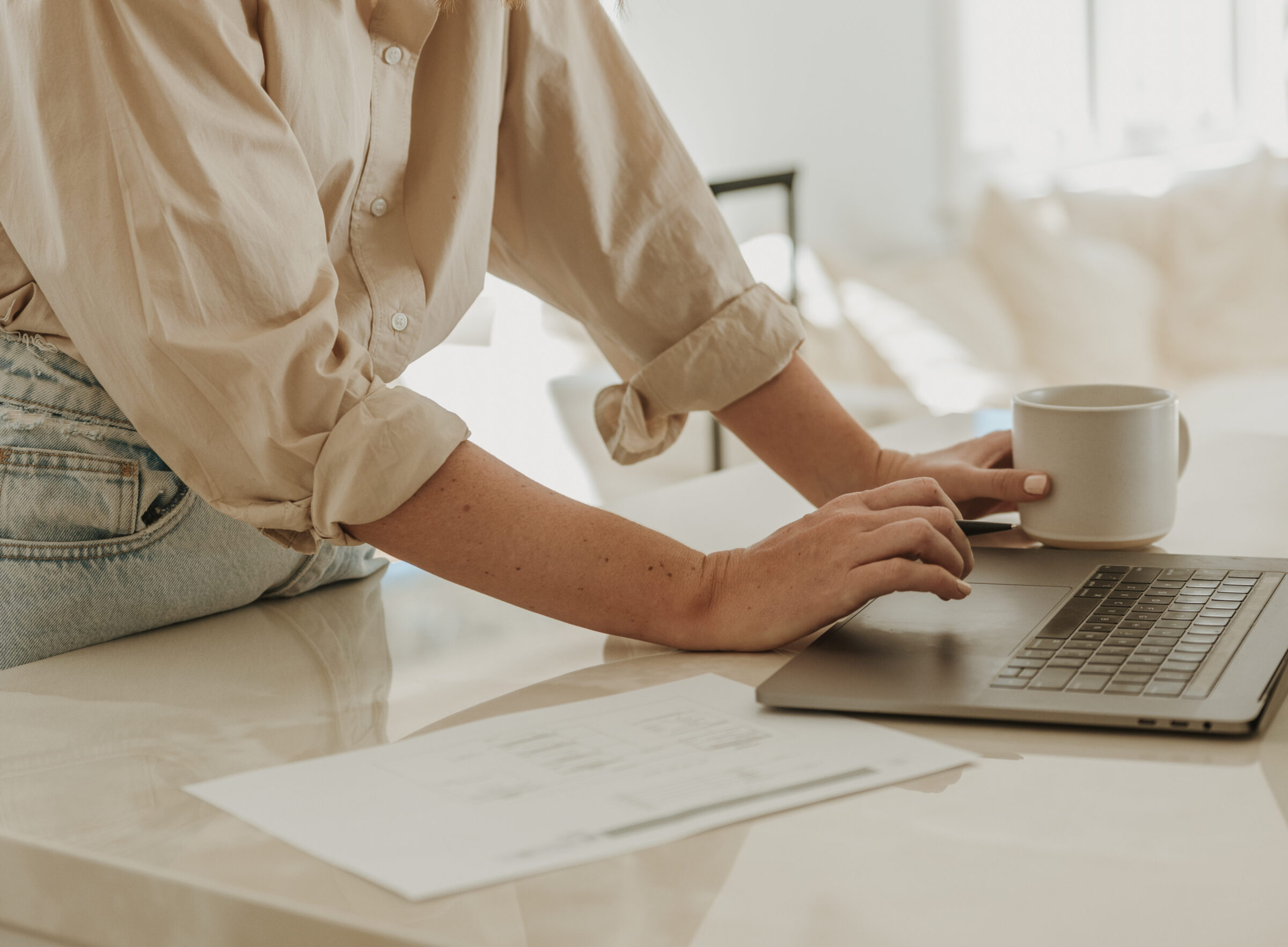 Woman at kitchen benchtop with a coffee mug, typing on a laptop beside a printed travel budgeting spreadsheet, demonstrating how to create a family travel budget effectively.