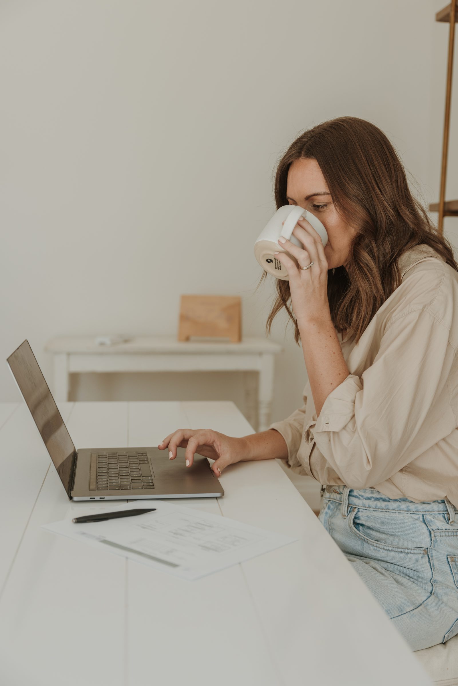 Mother taking a sip of coffee while working on her laptop at a desk, assembling a family vacation planner, illustrating the thoughtful process involved in how to plan for a family vacation, from budgeting to scheduling activities for everyone's enjoyment.