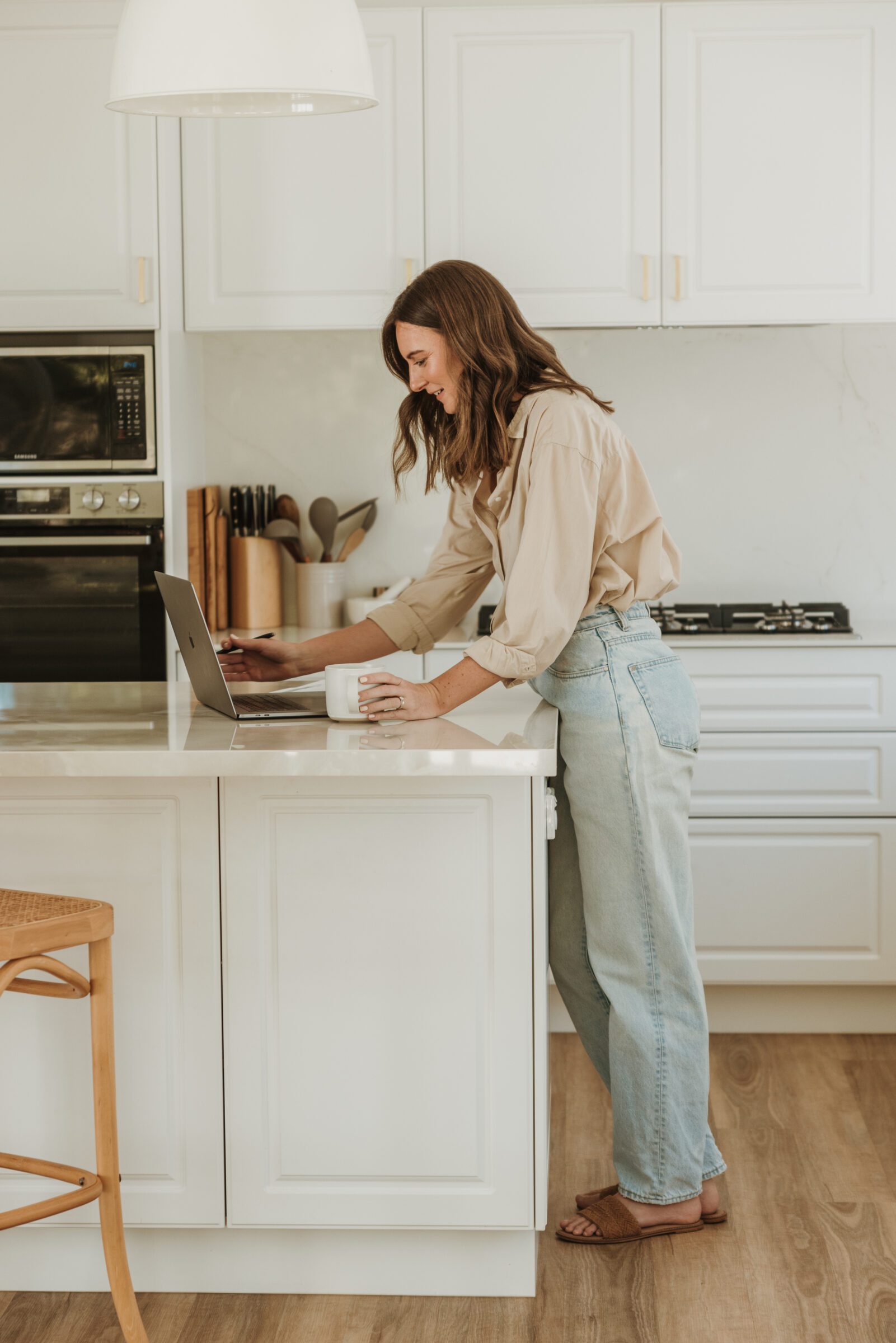 Woman standing at her kitchen bench with a coffee, typing on her laptop to establish her family's total travel budget on a spreadsheet, illustrating the planning phase of the high-low travel method for a balanced vacation experience.