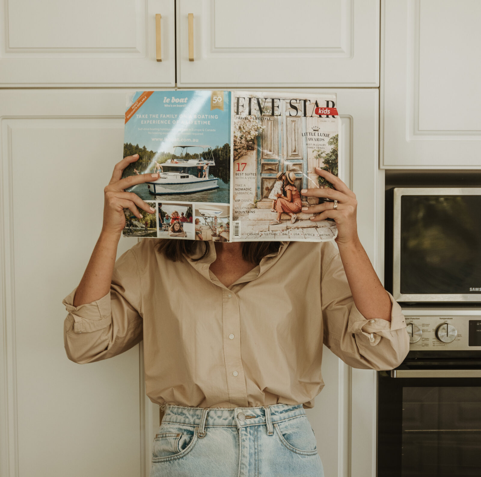 Woman in kitchen holding a 5-star travel magazine in front of her face, embodying the high-low travel approach by blending luxury aspirations with everyday settings.