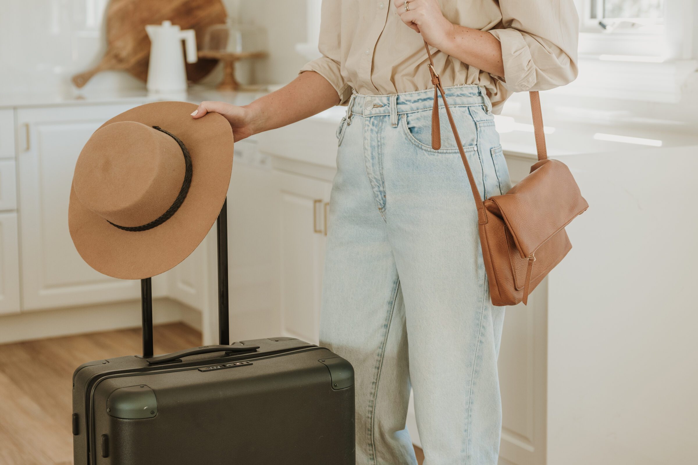 Woman ready to leave for the airport, standing next to her dark green suitcase with a brown handbag over her shoulder and a hat in one hand, epitomizing the best way to bring money on a trip by being prepared and organized.