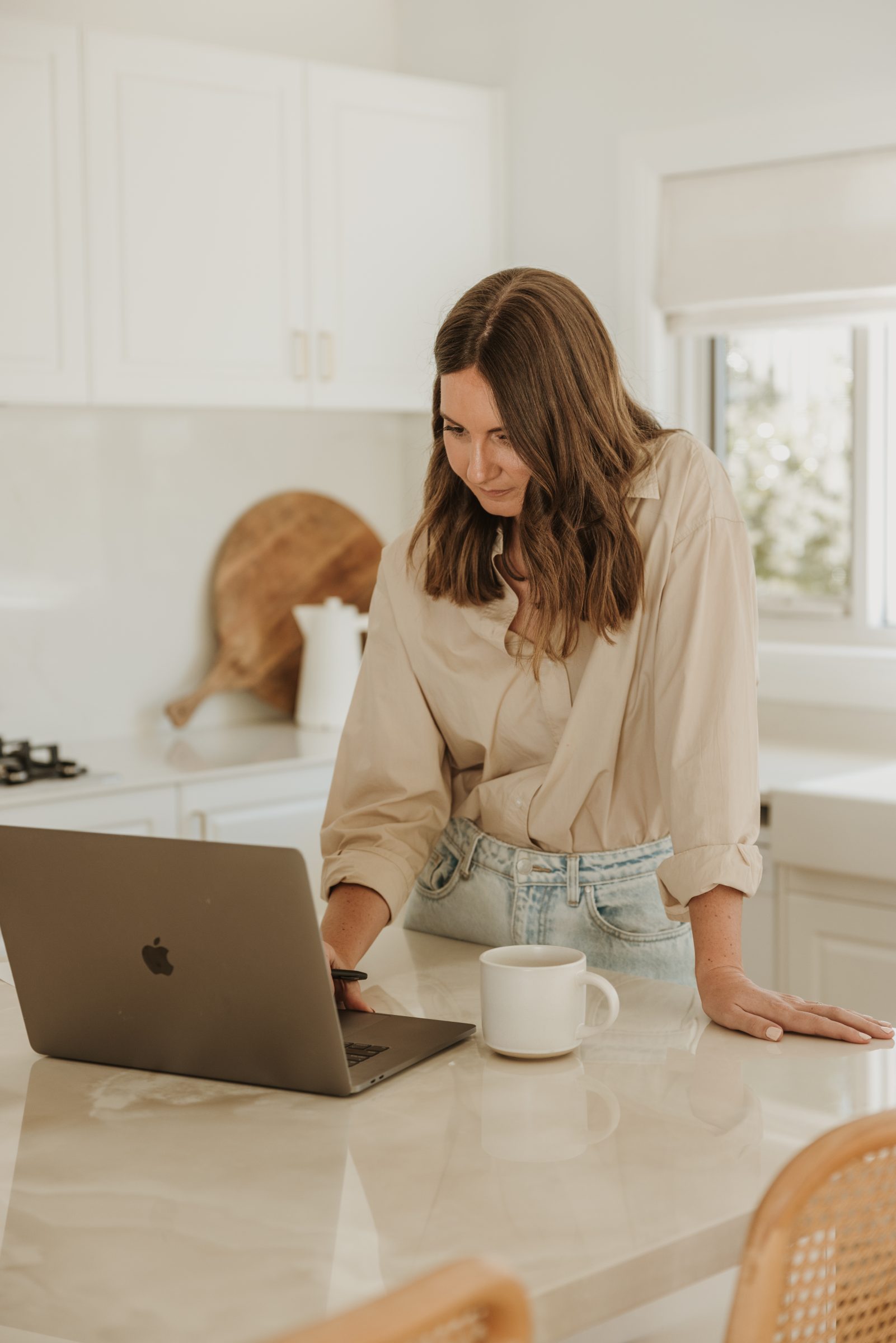 Woman standing at her kitchen bench, typing on a laptop with a pen in hand and a coffee mug beside her, organizing her finances for an upcoming family trip overseas, highlighting the preparation needed to manage money effectively while traveling.