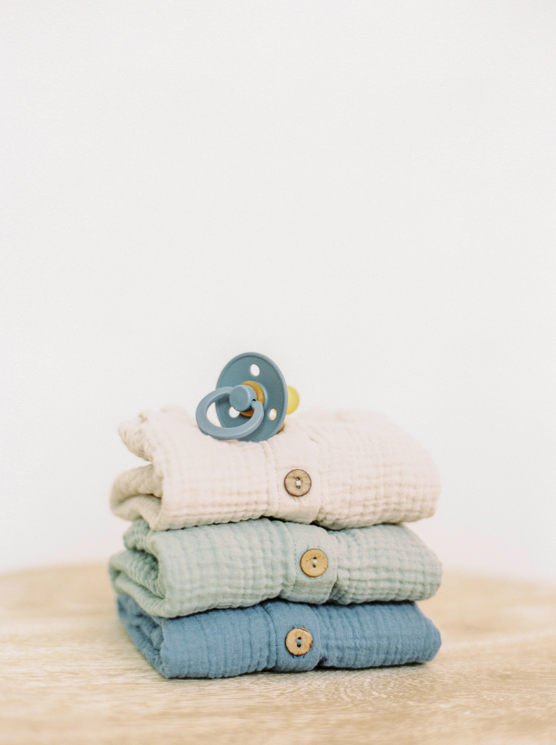 Folded baby clothing, including three short-sleeved rompers in blue, green, and white, with a blue pacifier on top, embodying the principle of 'how do you pack for travel with kids' by packing light for happiness on the go. Reminds parents that shops at most destinations can cover unexpected needs, easing travel with toddlers and babies.