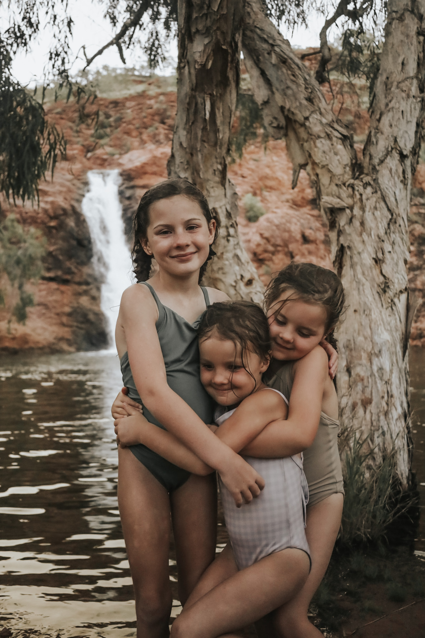 Three young girls standing in front of a natural waterhole in Kununurra with a waterfall in the background, captured during the rainy season, showcasing how selecting the travel season is a critical budgeting step. This image highlights the importance of timing in creating a family travel budget, as different seasons can significantly impact travel costs.