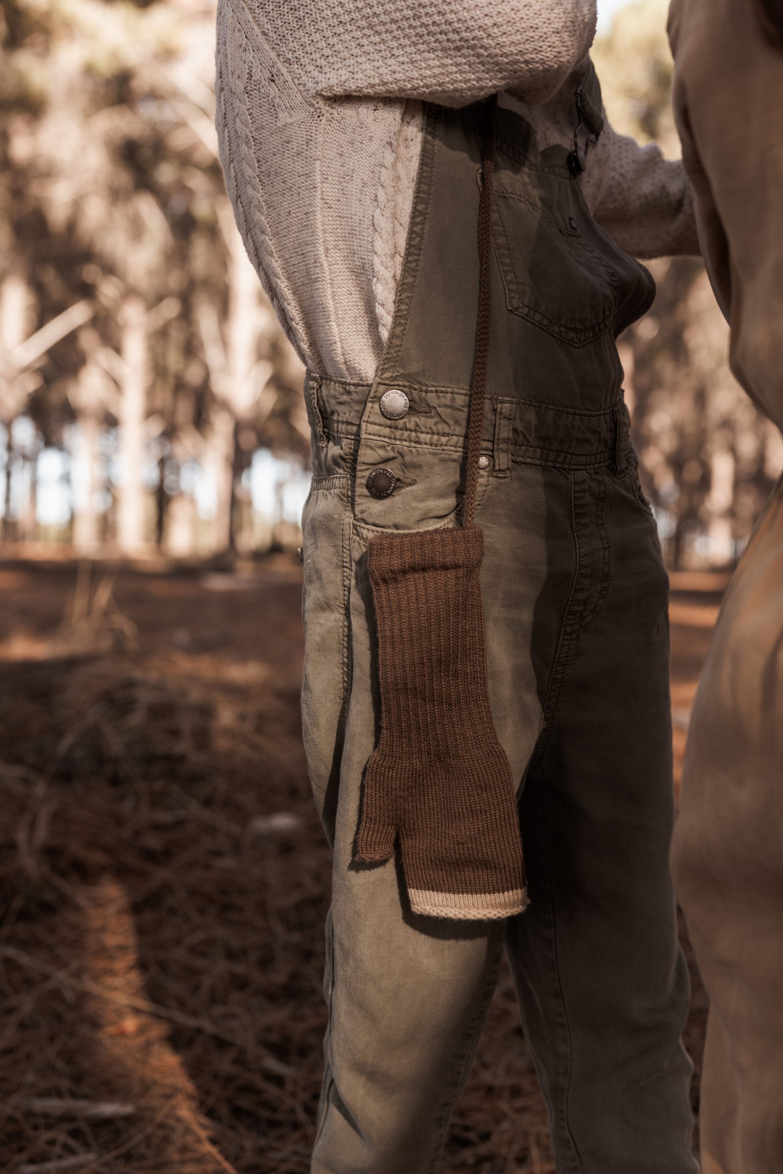 Close-up of a young girl in dark green overalls and a jumper, playing in the woods, with a focus on her glove, highlighting the essential tip on 'how do you pack for travel with kids'—remembering to pack for the weather, ensuring children are comfortably equipped for cold conditions.