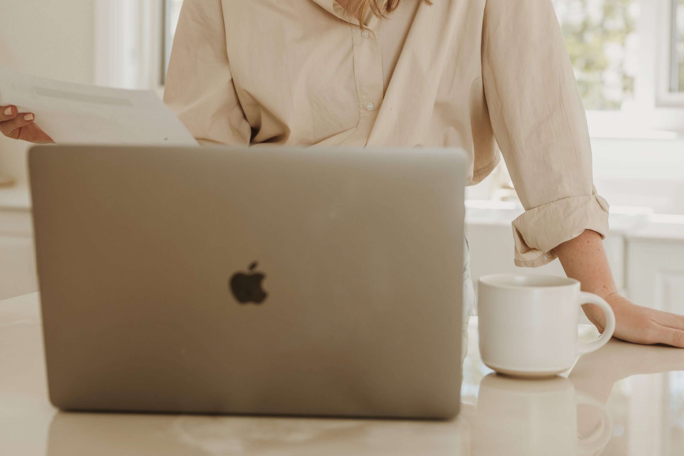 Woman at her kitchen island with a laptop and warm drink, reviewing a budgeting spreadsheet, demonstrating 'how to create a family travel budget' for an upcoming holiday, emphasizing financial planning for travel.