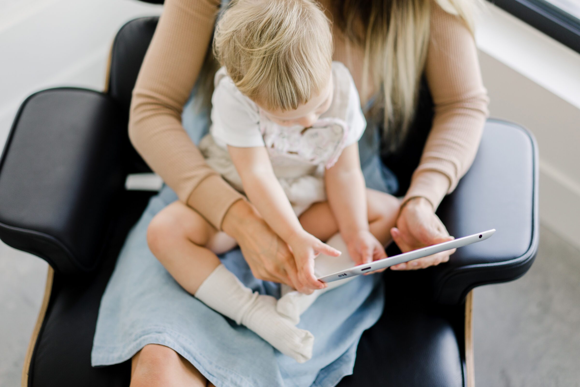 Mother sitting with her toddler on her lap, holding an iPad for him, capturing the balance of 'how do you pack for travel with kids' by incorporating electronics selectively. Illustrates electronics as both lifesavers during long journeys and something to use sparingly, ensuring screen time doesn't dominate the trip.