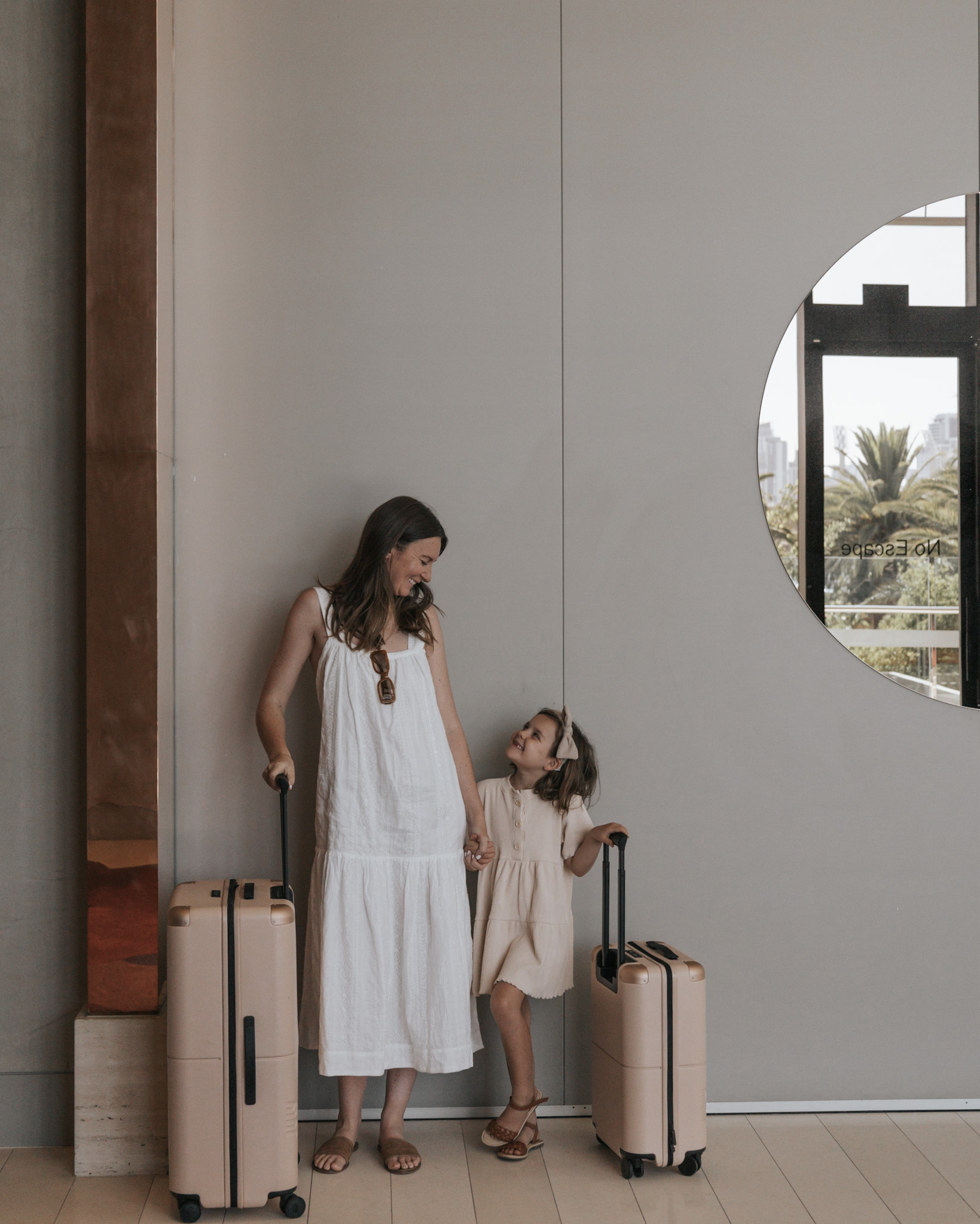 Mother and daughter each with a suitcase and backpack, smiling at each other in a hotel lobby, illustrating the ease of keeping luggage simple when traveling with kids. This image emphasizes the strategy of one backpack and one suitcase per person as ideal, capturing the essence of how do you pack for travel with kids.