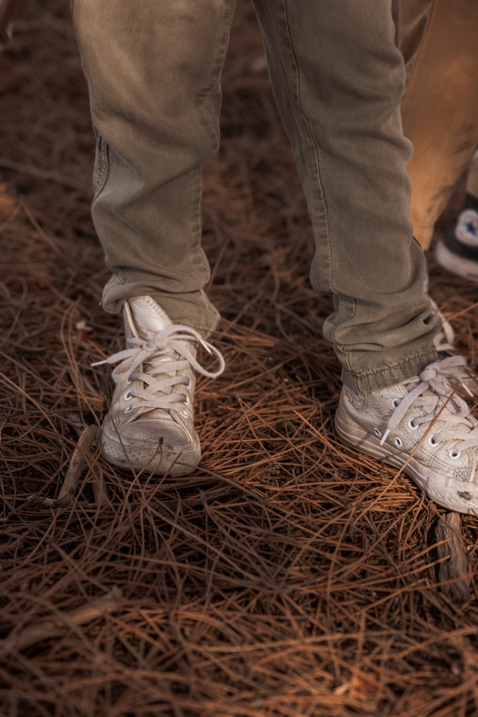 Close-up of a young girl in dark green overalls playing in the woods, with a focus on her white Converse shoes, illustrating the importance of selecting appropriate footwear when considering 'how do you pack for travel with kids'—ensuring comfort and suitability for various activities.