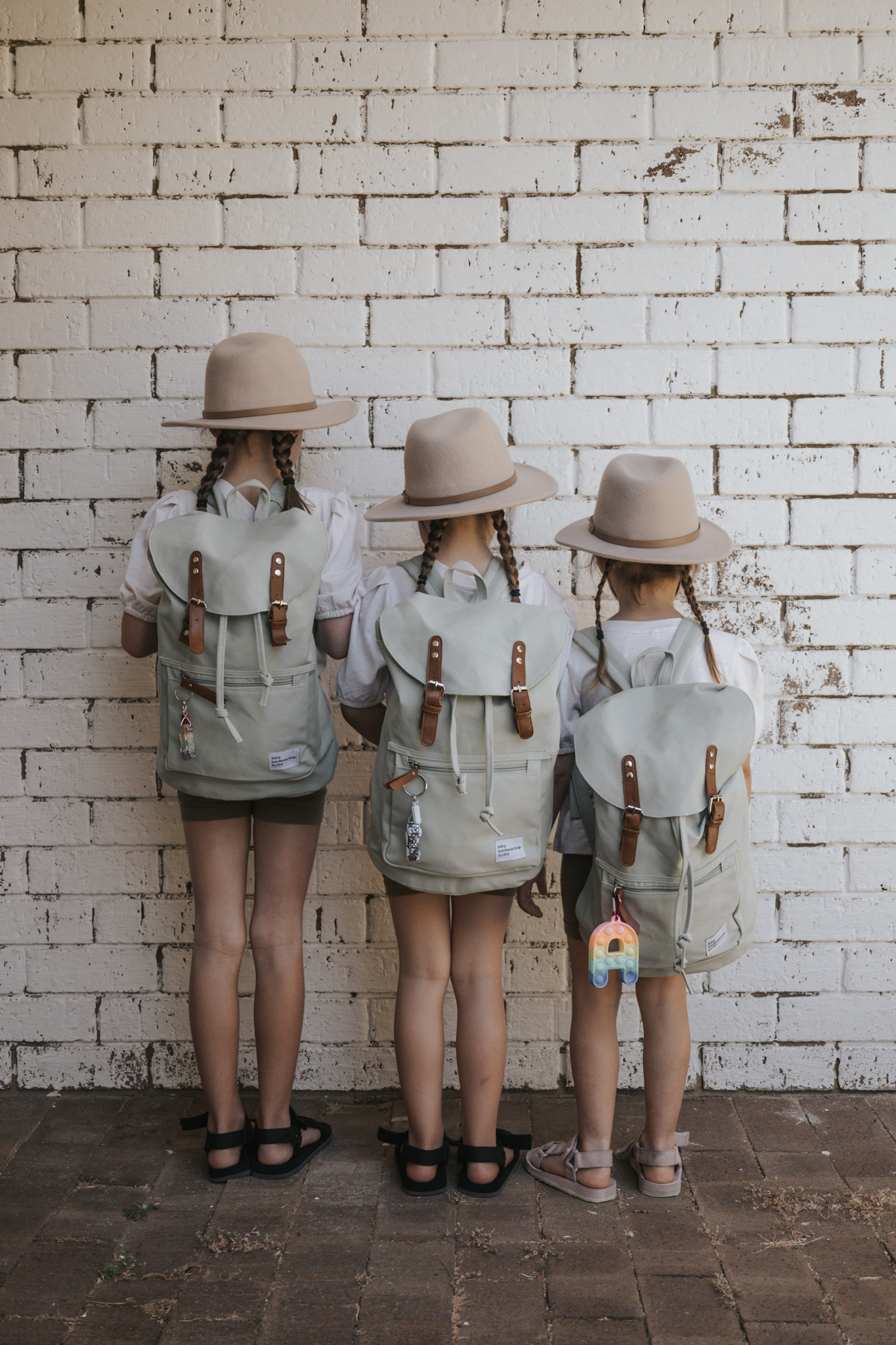 Three young sisters facing away from the camera, ready for travel with their green backpacks and hats, exemplifying how involving kids in the packing process lightens the load and empowers them, embodying the practical approach to 'how do you pack for travel with kids' for a smoother family travel experience.