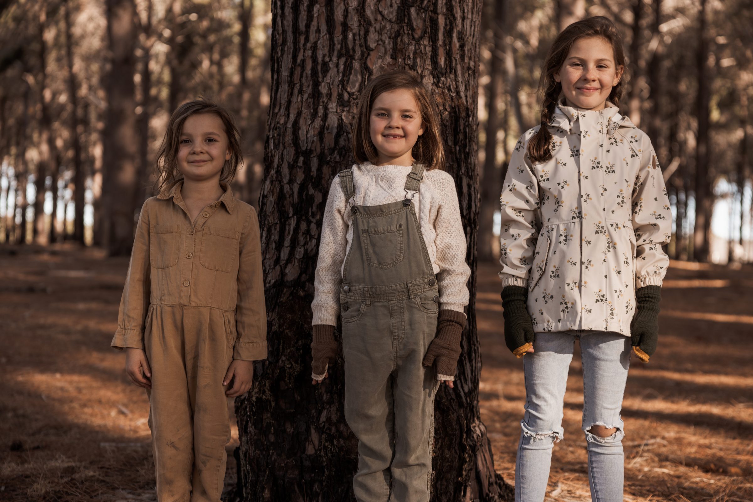 Three young sisters having fun in the woods, dressed in neutral-colored, hard-wearing clothing ideal for travel, showcasing the practicality of selecting durable and versatile outfits when considering how do you pack for travel with kids, ensuring comfort and suitability for adventures.