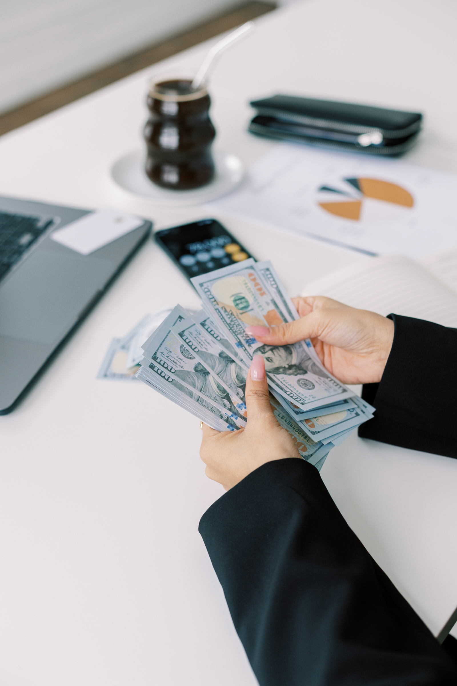 Woman at desk holding cash notes, surrounded by essentials like a laptop and coffee, illustrating the concept of allocating weekly spending money. By budgeting a fixed amount for discretionary spending each week, individuals gain insights into their spending habits, fostering financial mindfulness and saving towards travel goals.