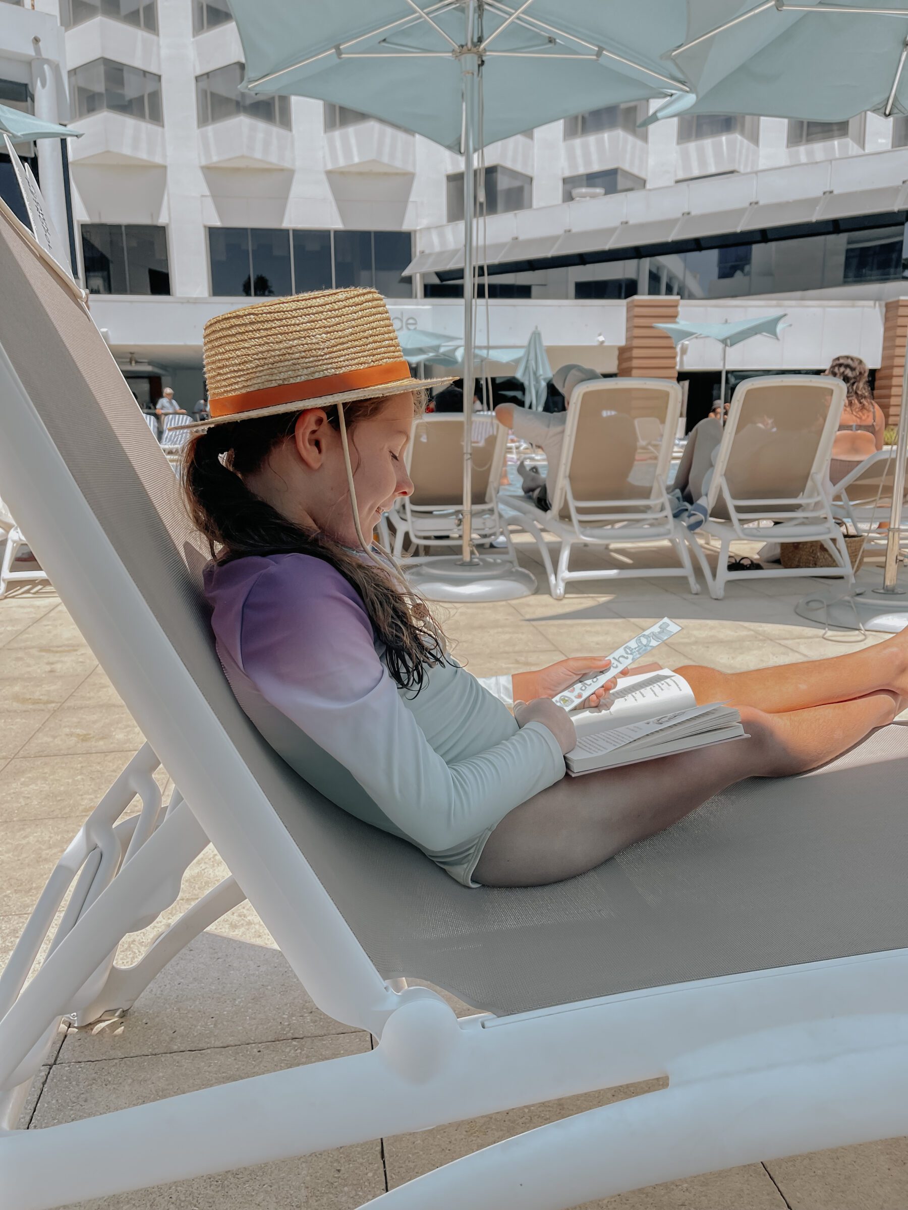 Young girl sitting on a pool chair at a hotel, dressed in bathers and a hat, reading a book. This image highlights the joys of a staycation, demonstrating that memorable holidays don’t require distant travel, and showcases how local hotels or Airbnb stays can offer refreshing new perspectives and support local businesses.