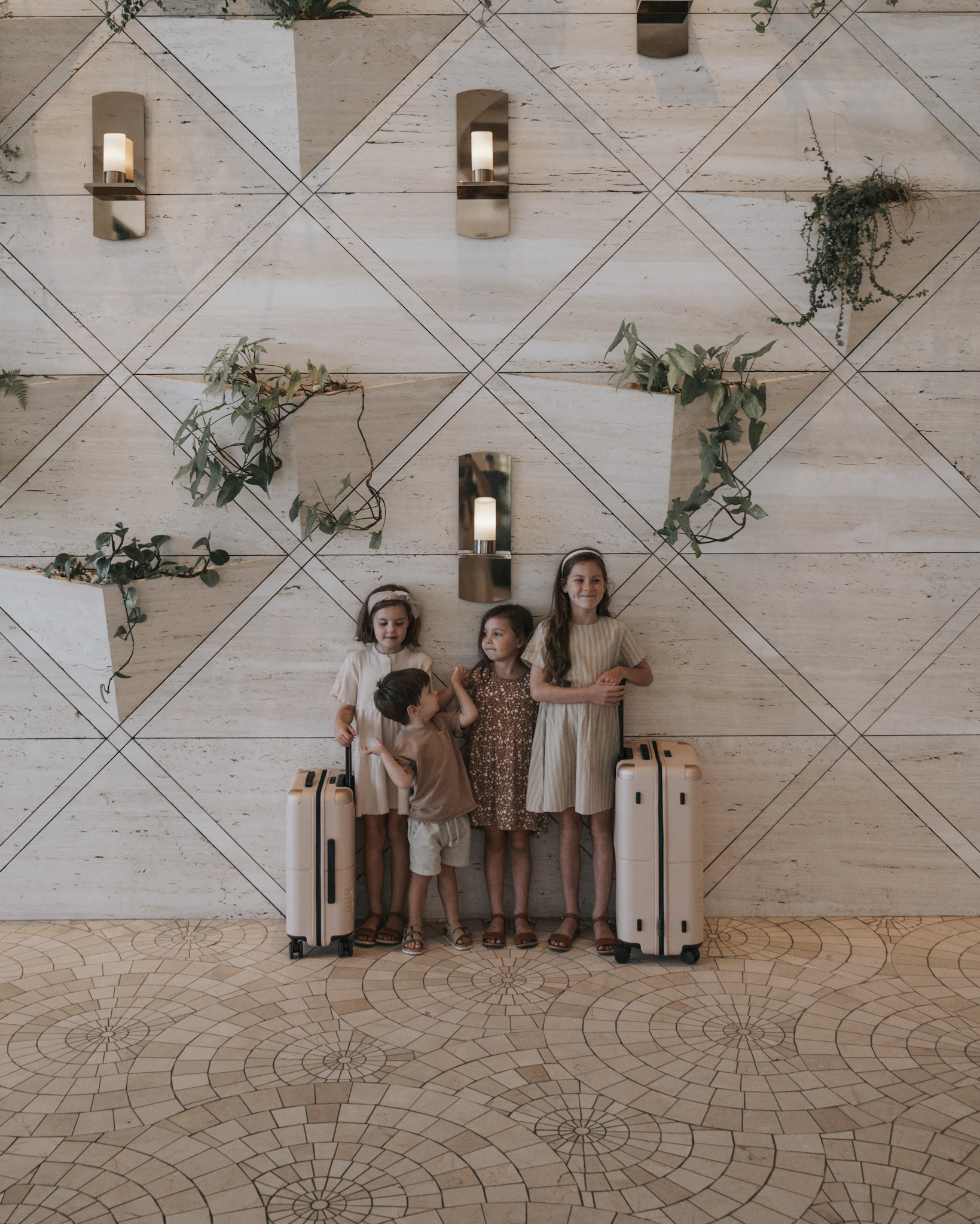 Four young siblings standing next to their suitcases at a beautiful hotel, demonstrating how to afford your next family vacation by booking flights and accommodation early. This image underscores the benefits of early planning, which secures lower prices and better choices due to supply and demand, easing financial pressures by allowing more time to save.