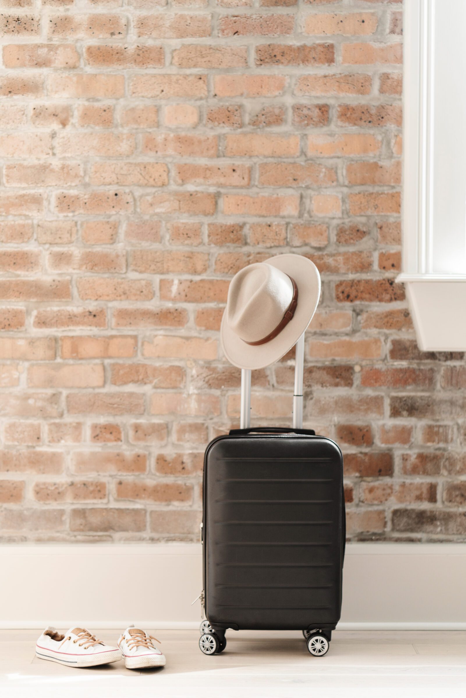 Black carry-on suitcase with a wide-brimmed hat and white sneakers, set against an exposed brick wall. Traveling carry-on only saves on checked baggage fees and simplifies airport navigation, ideal for family trips that don't require bulky gear.