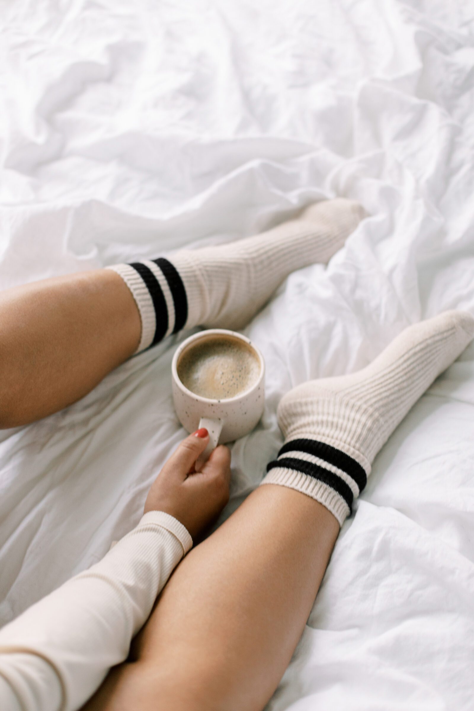 Woman sitting on her white bed, wearing comfy socks and holding a coffee, illustrating a simple way to afford your next family vacation by reducing daily expenses. This image shows how making coffee at home instead of buying it can help save money for travel, emphasizing practical budgeting tips for vacation savings.