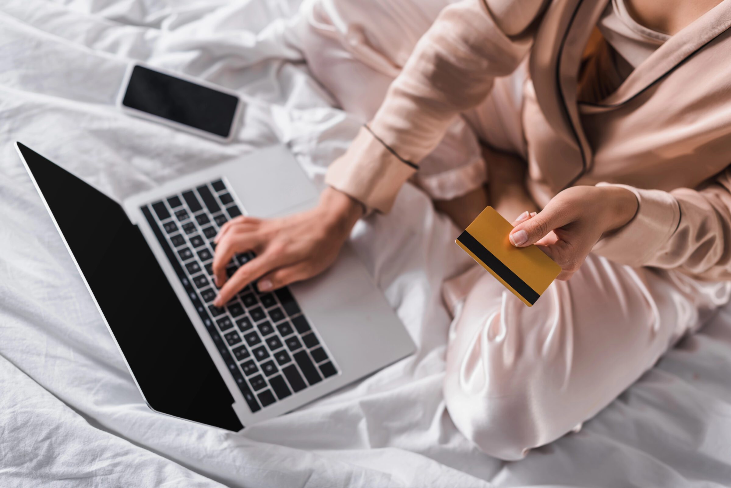 Woman sitting in bed with smartphone, credit card, and laptop, checking card limits and capacities as part of preparing a secure travel money system overseas.