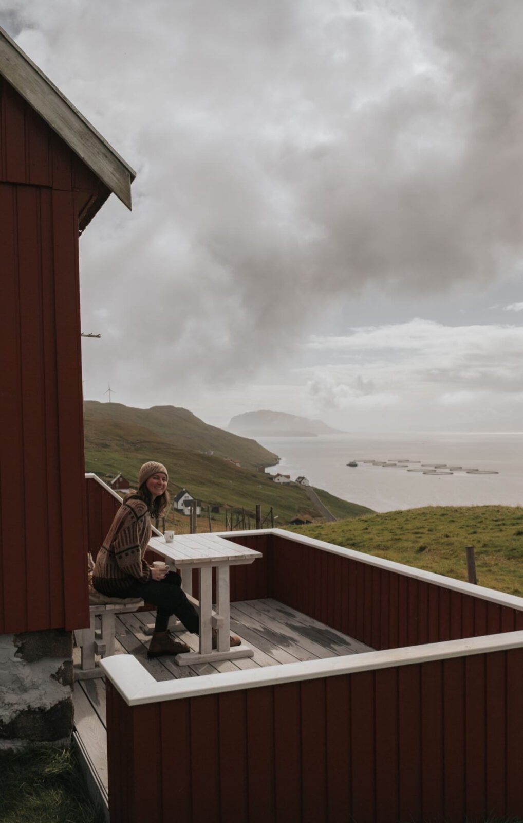 Woman sitting on the front balcony of a cabin in the Faroe Islands, overlooking the ocean and hills, representing using tax returns and windfalls to boost travel savings.