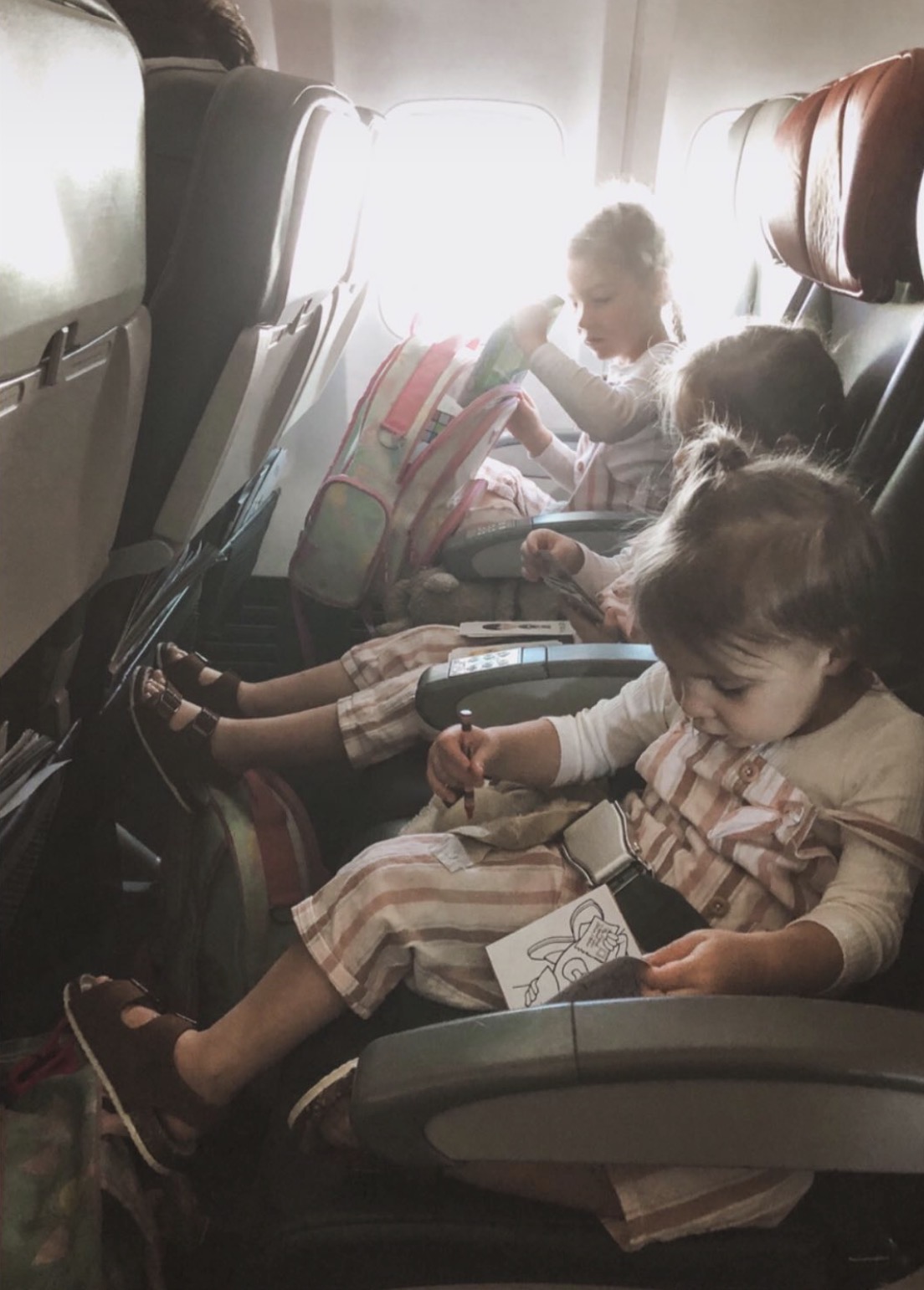 Three sisters in matching striped outfits sit buckled into a row of plane seats, colouring in and snacking with a pink backpack tucked by the window.