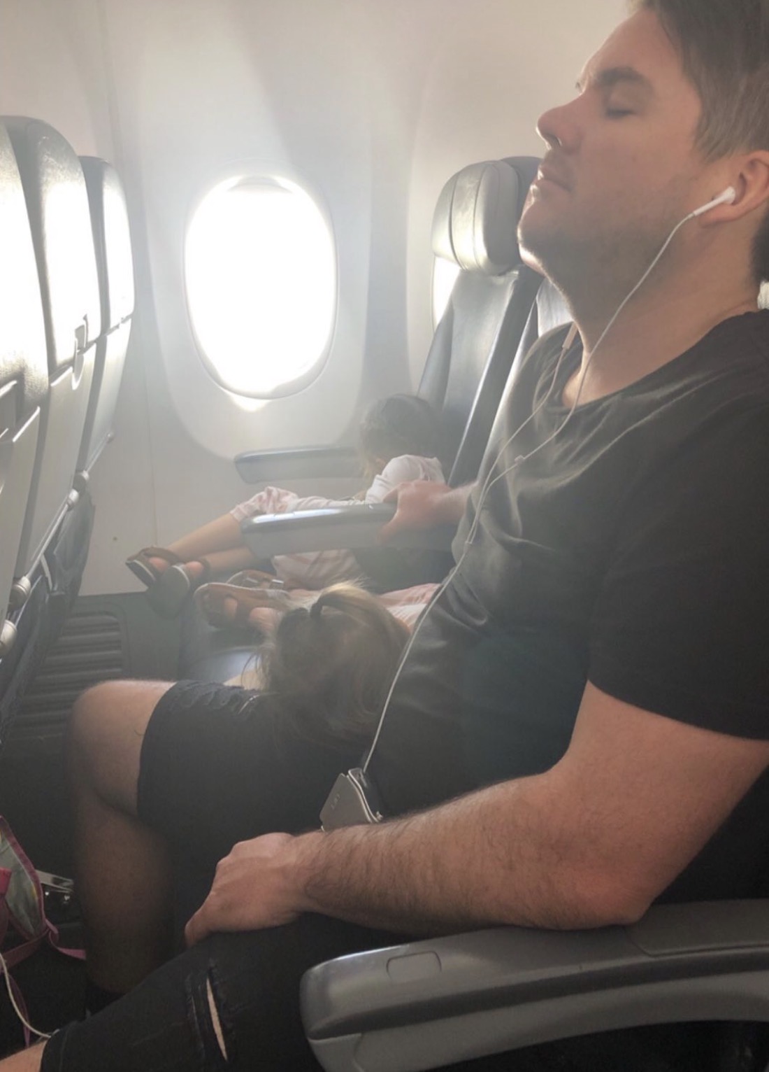 A dad naps with earbuds in as a toddler girl sleeps curled up across his lap, sunlight flooding through the cabin windows behind them.
