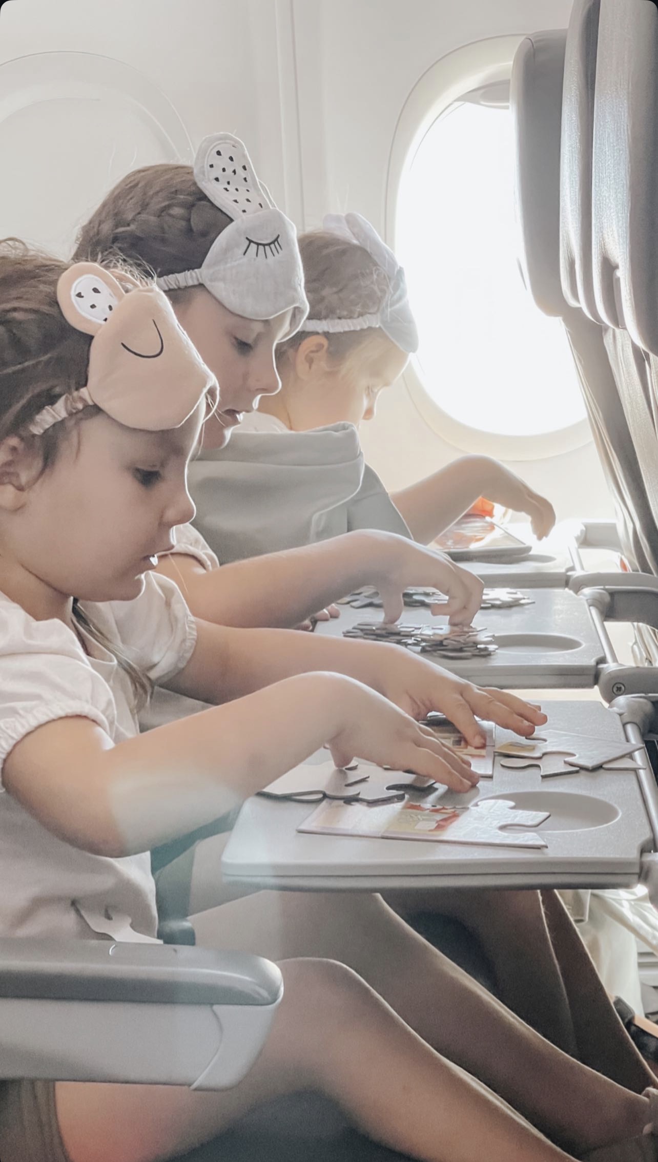 Three young girls in animal-ear eye masks sit in a row of plane seats, absorbed in putting together puzzle pieces spread across their tray tables in the sunlight.
