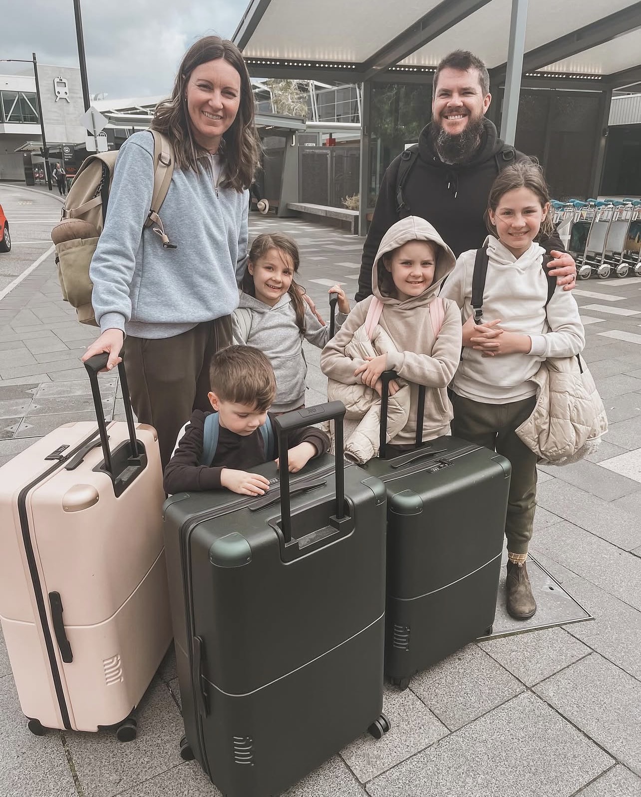 A mum and dad pose with their four kids outside the airport, all smiling behind a line-up of matching blush pink and khaki suitcases, packed and ready to fly.