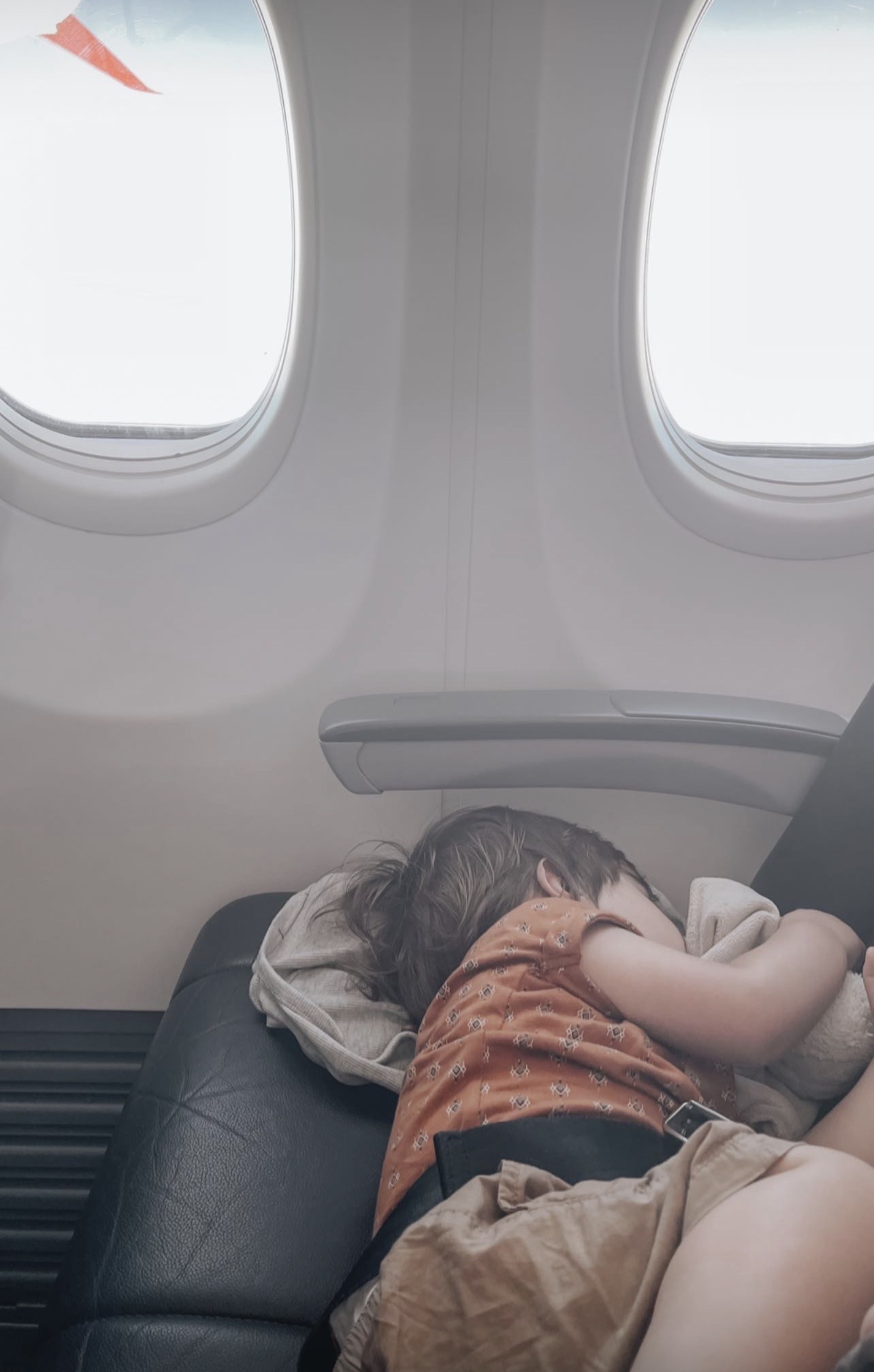 A toddler sprawls sideways across the seat fast asleep, cheek squished into a soft blanket, with two sunlit windows glowing above.